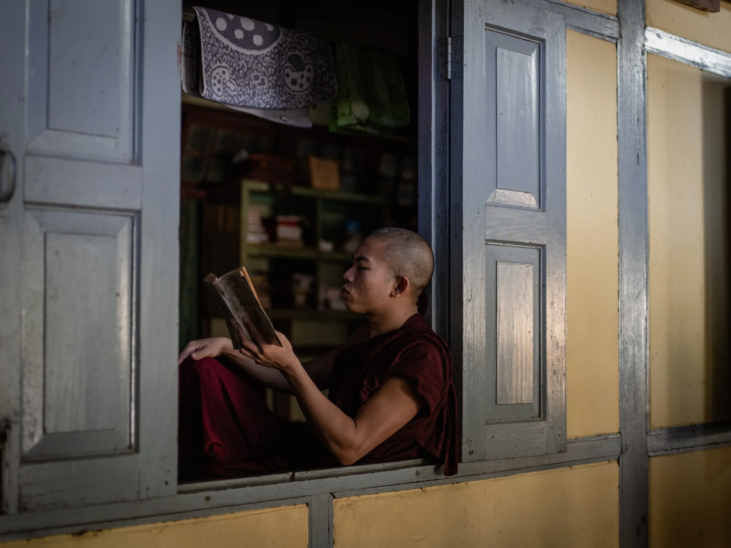 Young Buddhist monk in red robes reads a book while seated in an open wooden window.