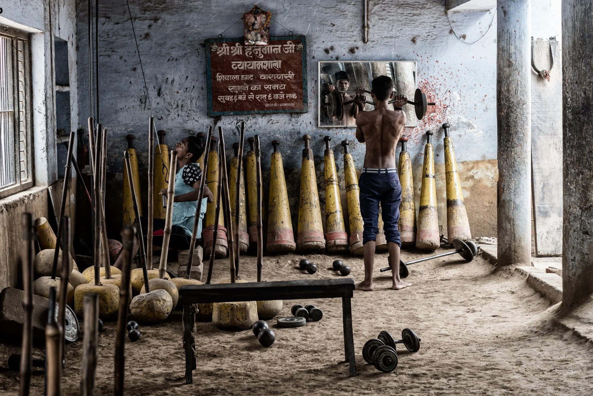 Young athlete trains with weights inside a traditional Indian gym, surrounded by wooden clubs, dumbbells, barbells, and Hindi signage.