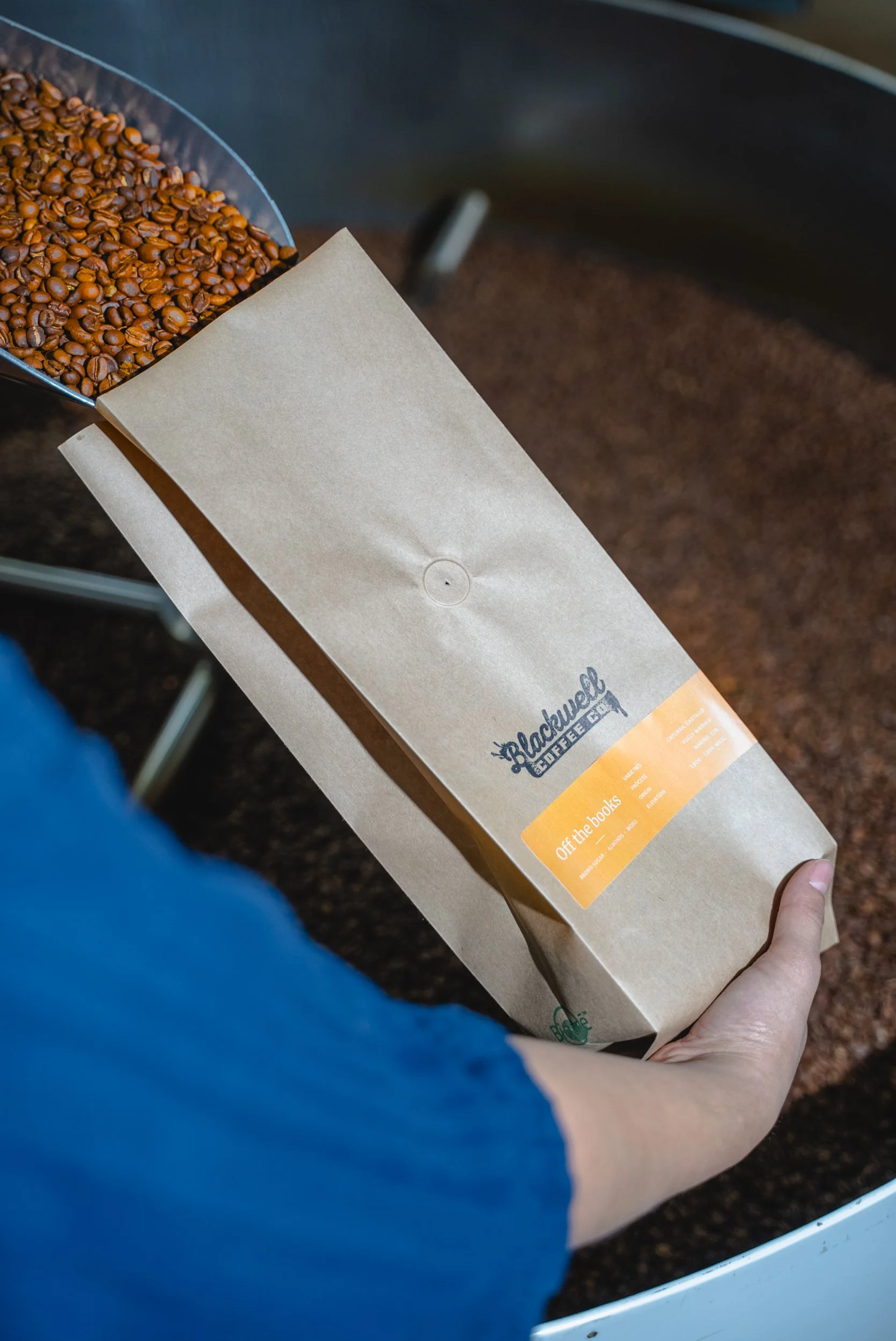 A person holding a paper bag of black coffee beans at a coffee shop. The bag has a label with the Blackwell Coffee logo and some text.