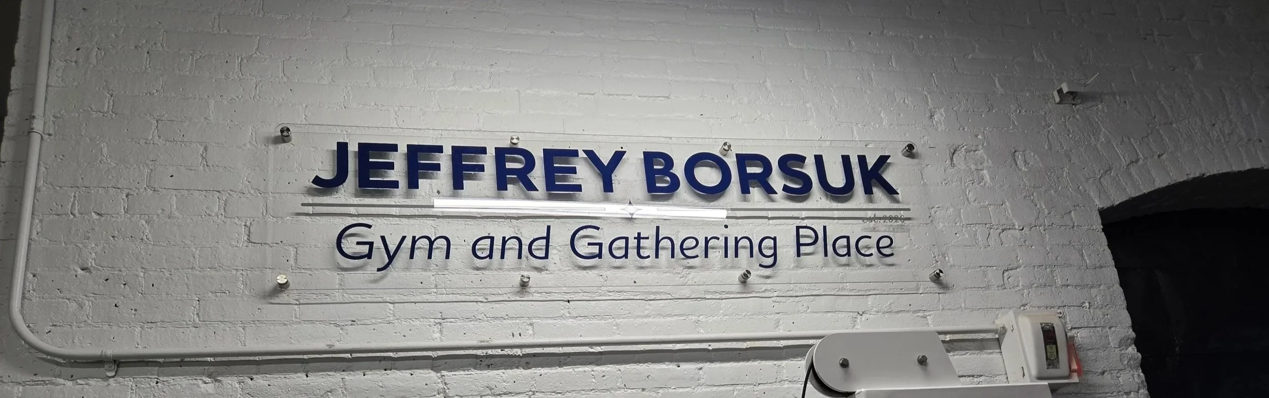 Jeffrey Borsuk Gym & Gathering Place Written in Blue Ink on Clear Plexiglass hanging on a white brick wall