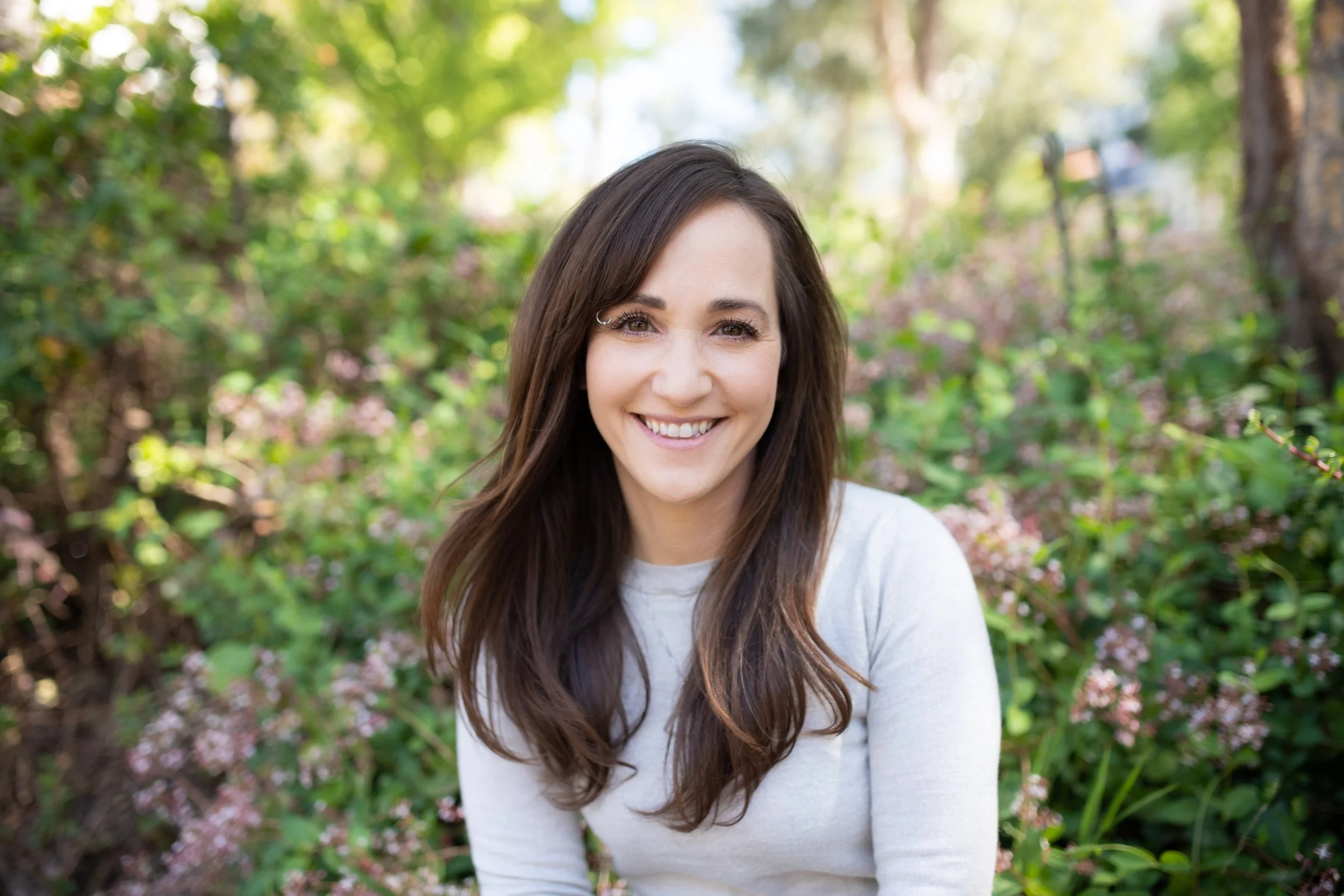 A woman with long brown hair smiling outdoors in a garden with green foliage and pink flowers.