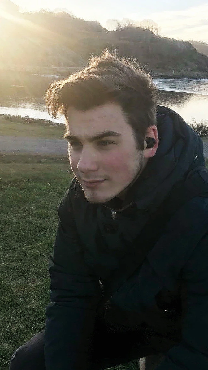 Young man outdoors near a body of water with hills in the background during sunset, wearing a black jacket and wireless earbuds.
