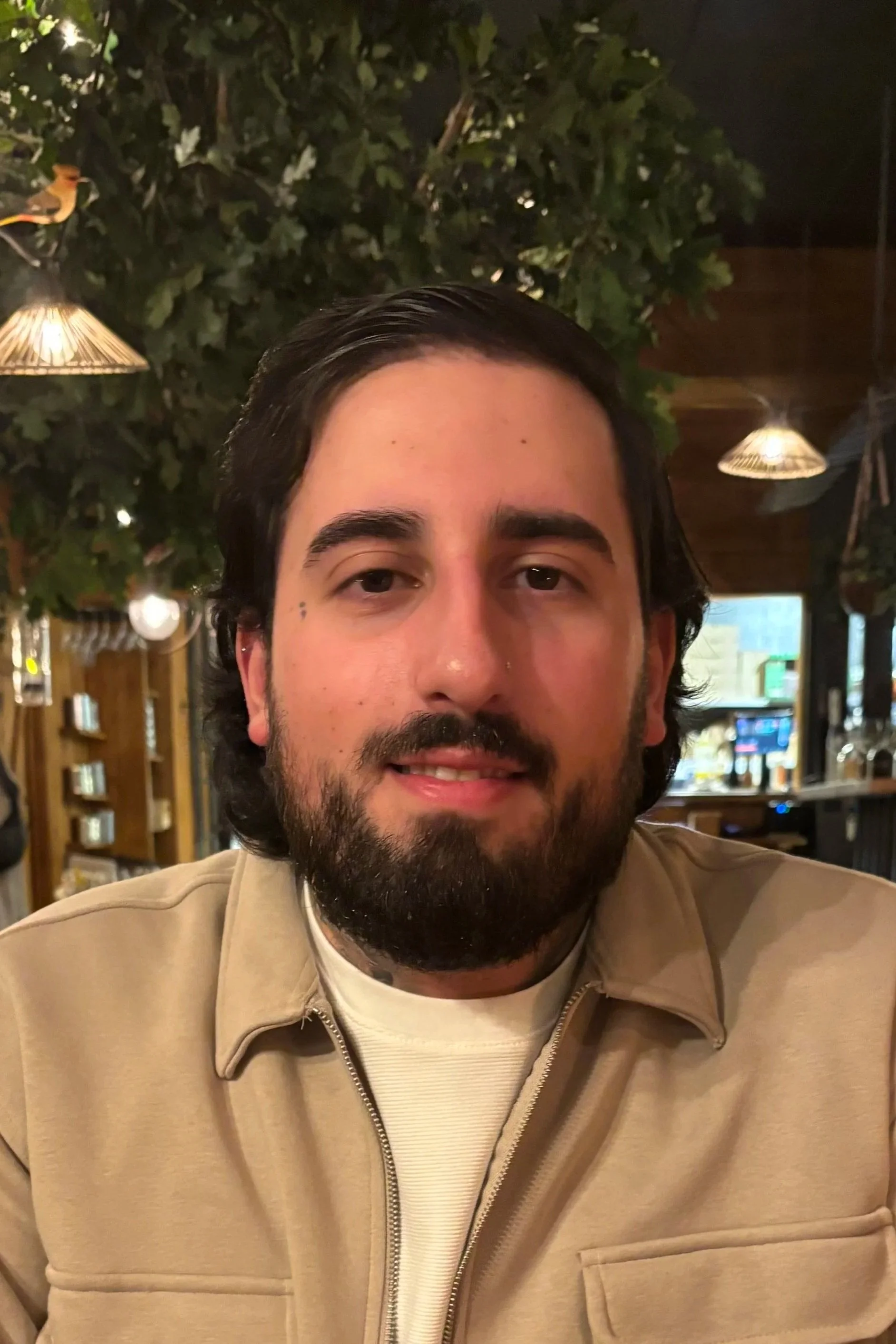 A man with dark hair and a beard sitting inside a restaurant or cafe, with hanging lights and greenery in the background.