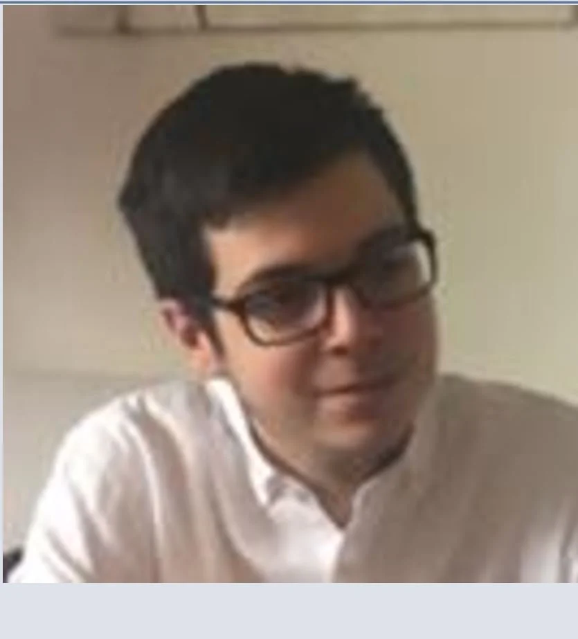 A young man with short dark hair and glasses, wearing a white collared shirt, smiling slightly in an indoor setting.