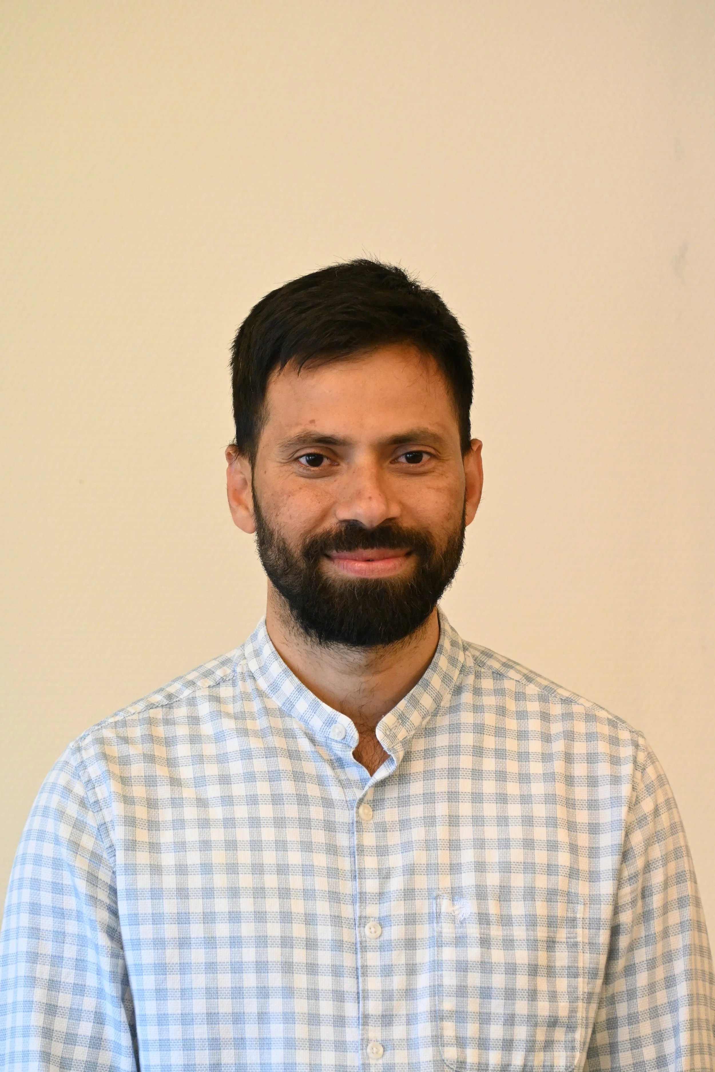 A man with dark hair and a beard, wearing a light-colored checkered shirt, standing against a plain beige background.