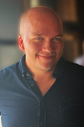 A bald man in a dark blue button-up shirt smiling at the camera, indoors with blurred background.