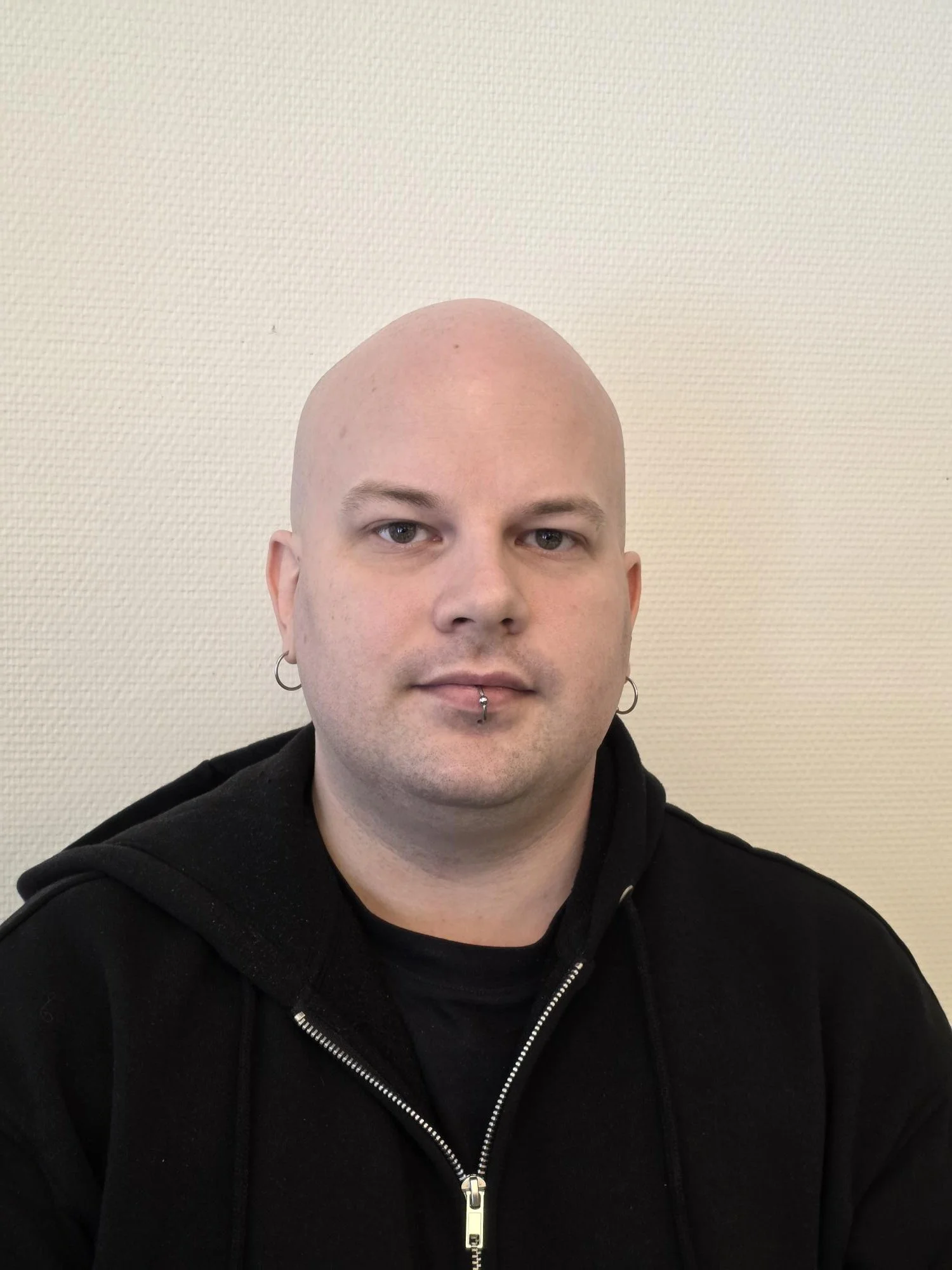 A bald man with earrings and a septum piercing wearing a black hoodie, sitting against a beige wall.