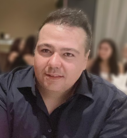 A man with short dark hair and light skin tone wearing a dark shirt, sitting at a table in a social setting with blurred people in the background.