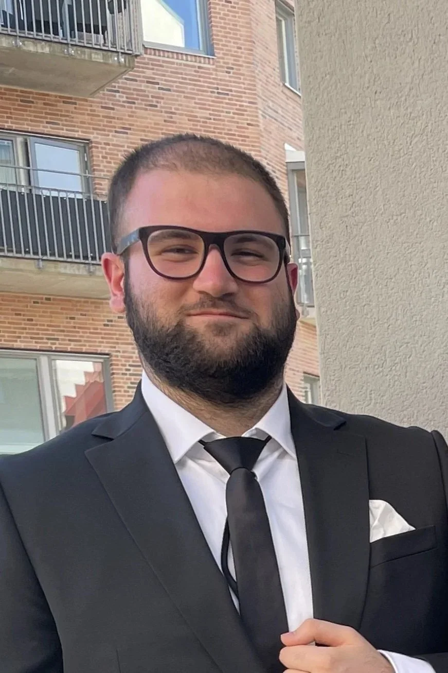 A man with glasses and a beard dressed in a black tuxedo with a white shirt and black tie, posing outdoors near a building with brick and beige walls.