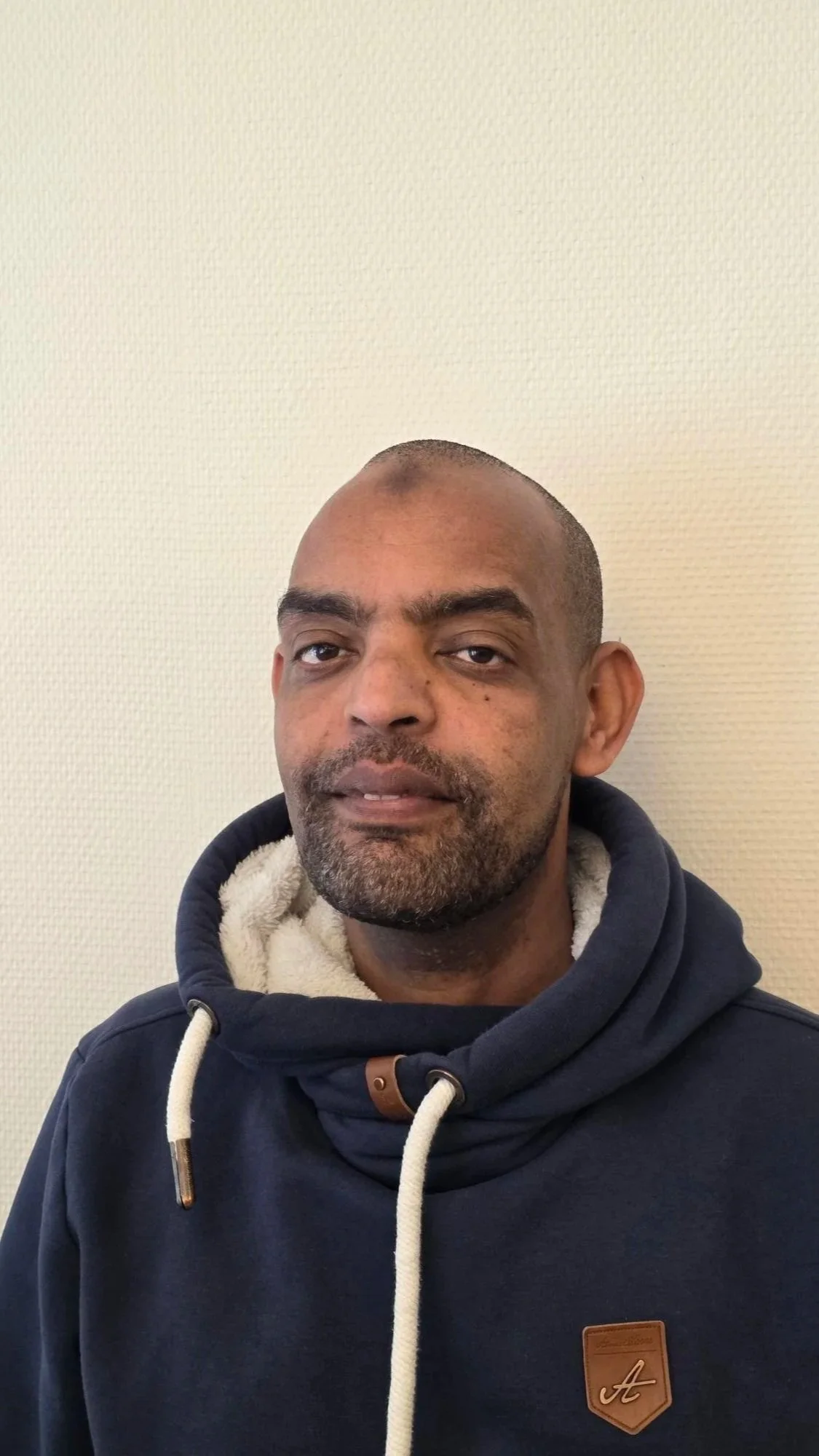 A man with a beard and short hair wearing a dark blue hoodie with tan accents in front of a plain, off-white wall.