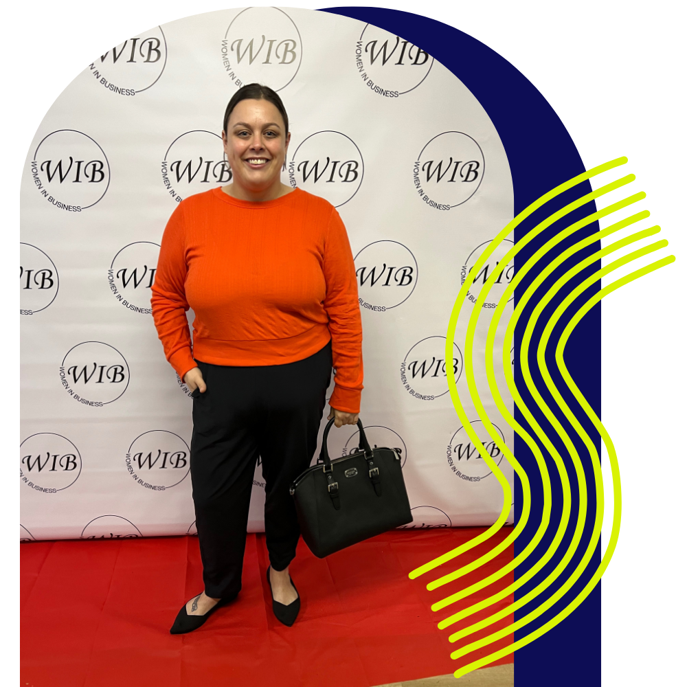 A woman standing on a red carpet in front of a backdrop with the Women in Business logo, wearing an orange sweater, black pants, and black shoes, holding a black handbag.