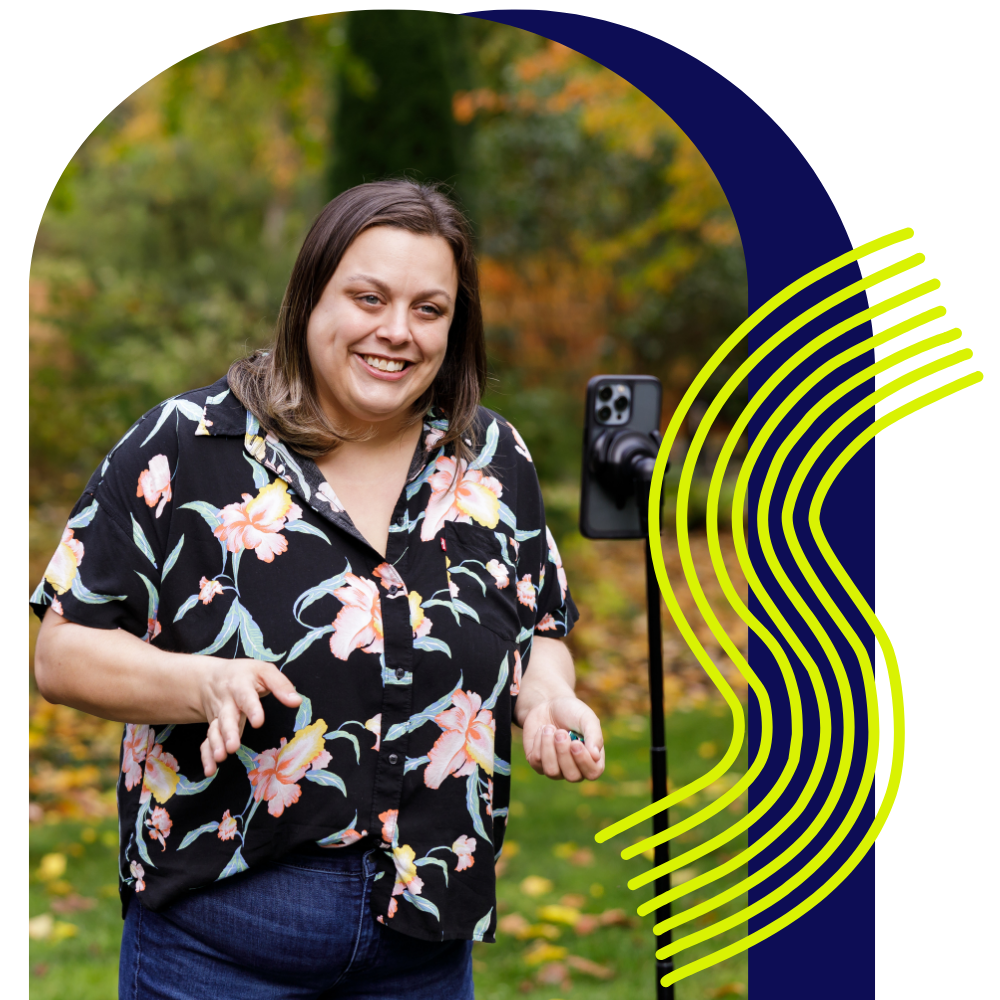 A woman smiling in front of a smartphone mounted on a stand outdoors in a park with autumn foliage.