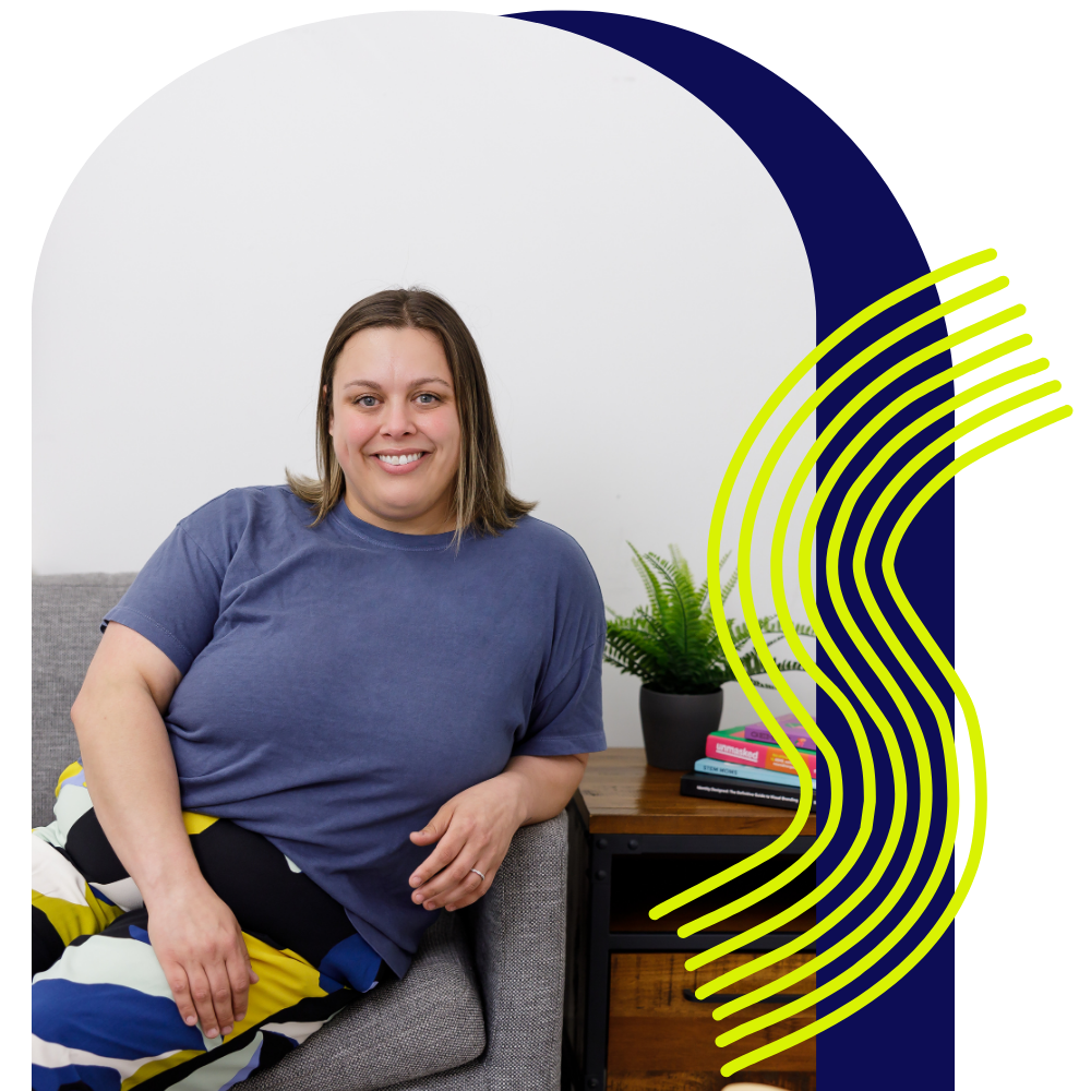 A woman with shoulder-length hair sitting on a gray couch, smiling, wearing a blue t-shirt and colorful pants, with a side table with books and a plant in the background.