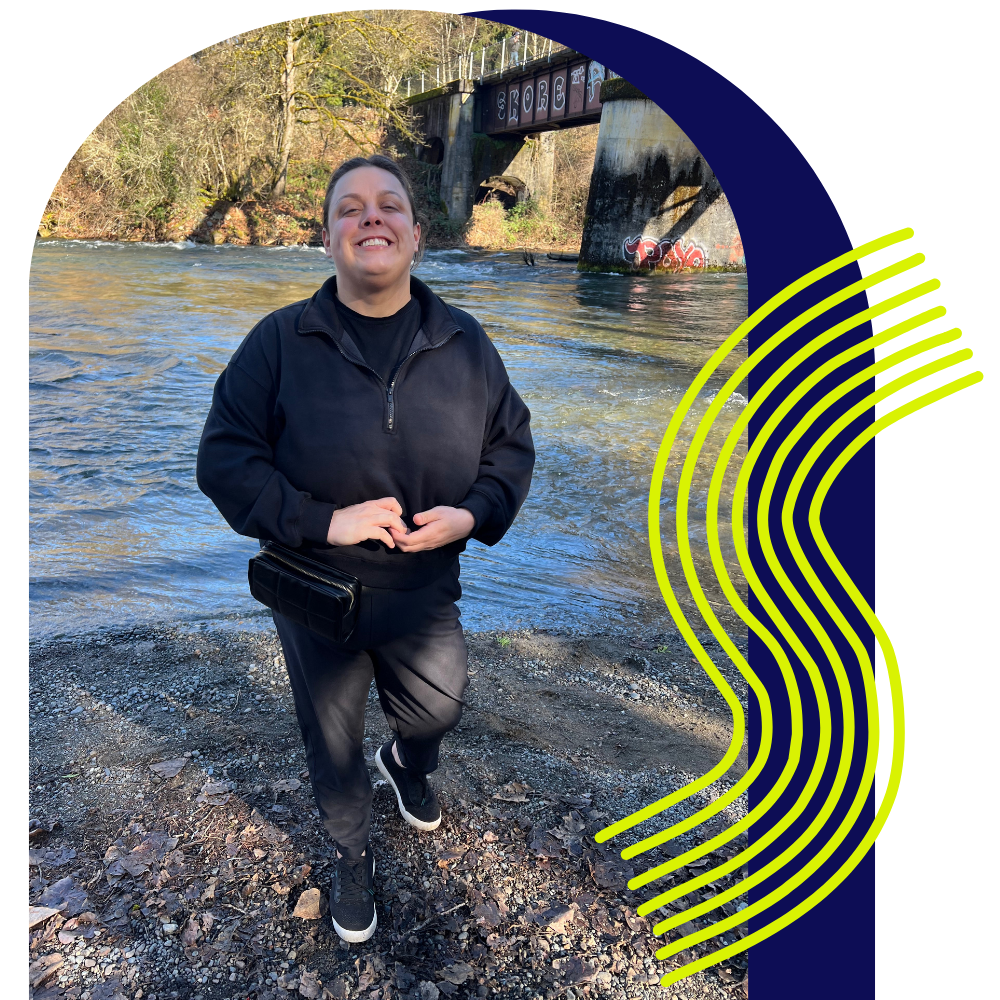 A woman standing by a river and smiling, wearing a black sweatshirt and pants, with trees and a graffiti-covered bridge in the background.