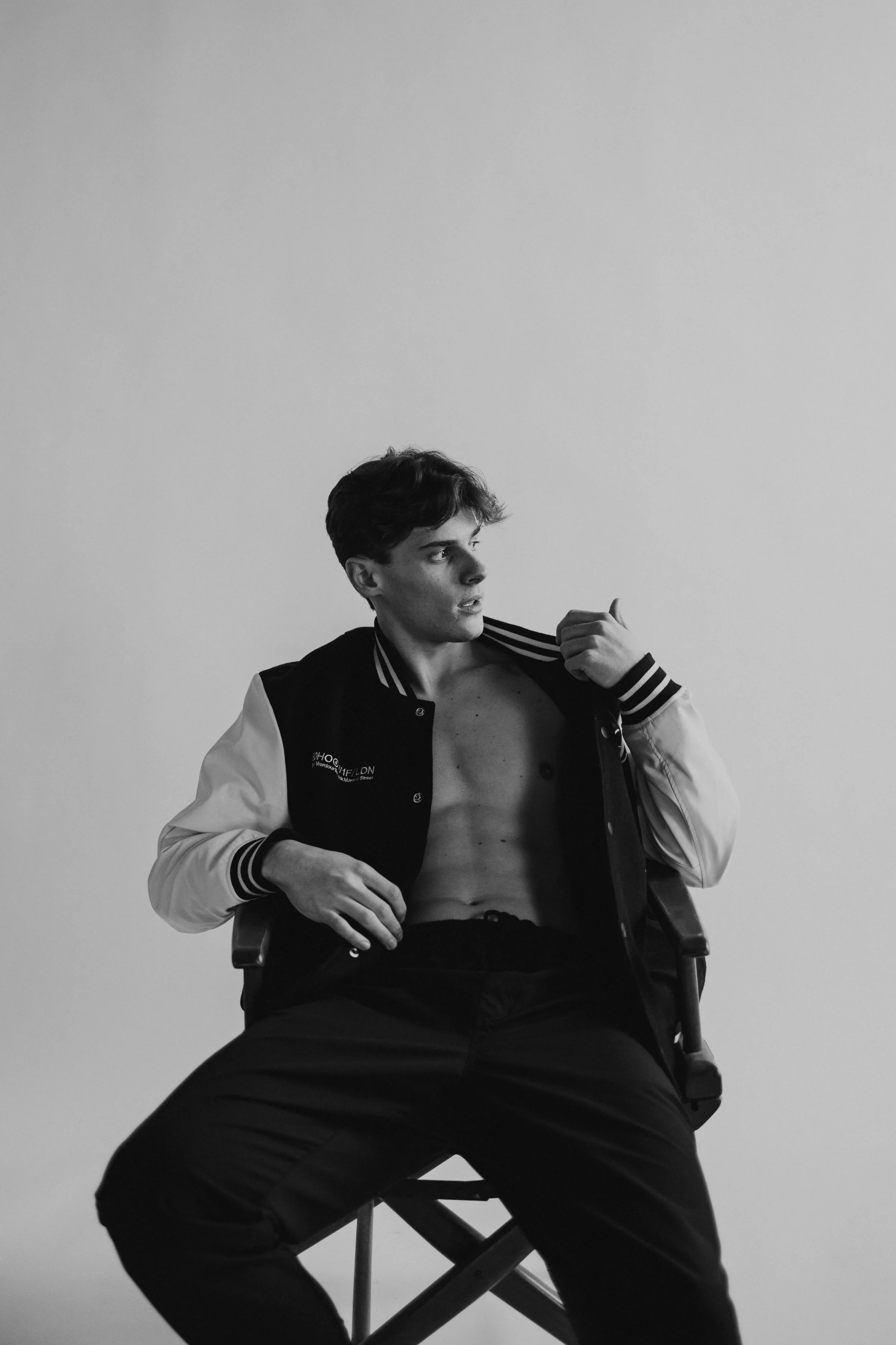 A young man sitting on a chair, shirtless, wearing a varsity jacket with a blank background.
