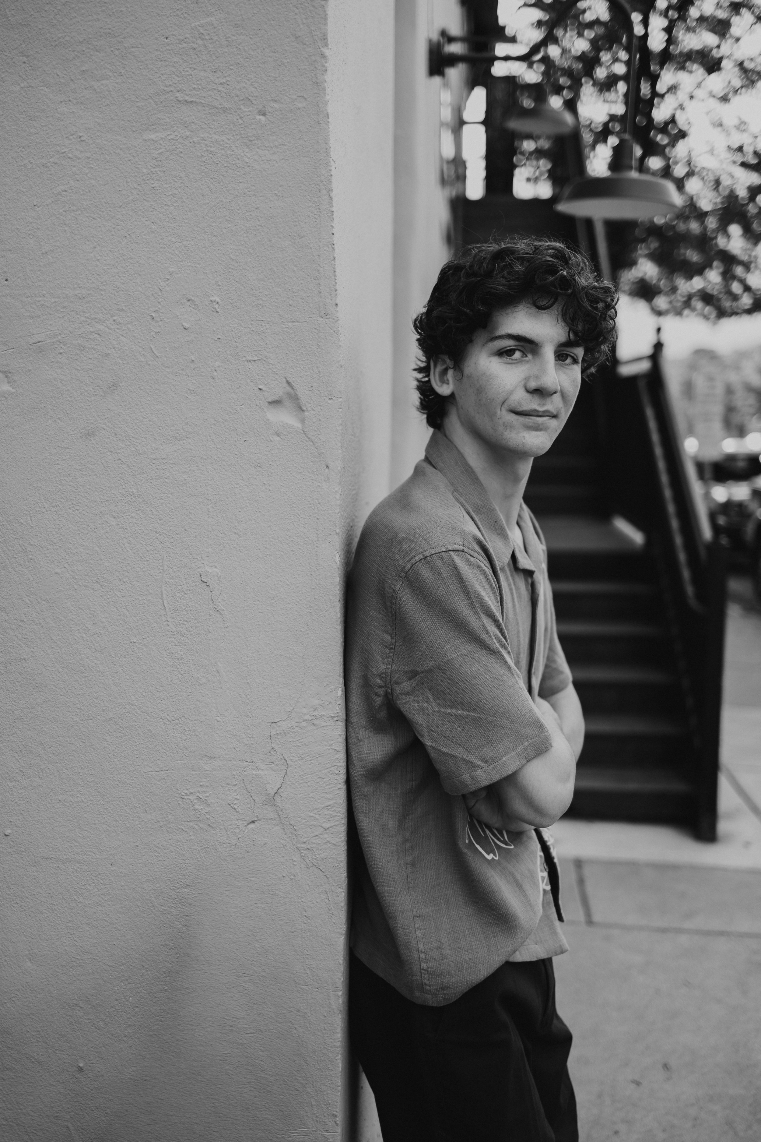 A young man with curly hair and a slight smile, dressed in a short-sleeved shirt, leaning against a wall on an urban sidewalk, with stairs and trees in the background.