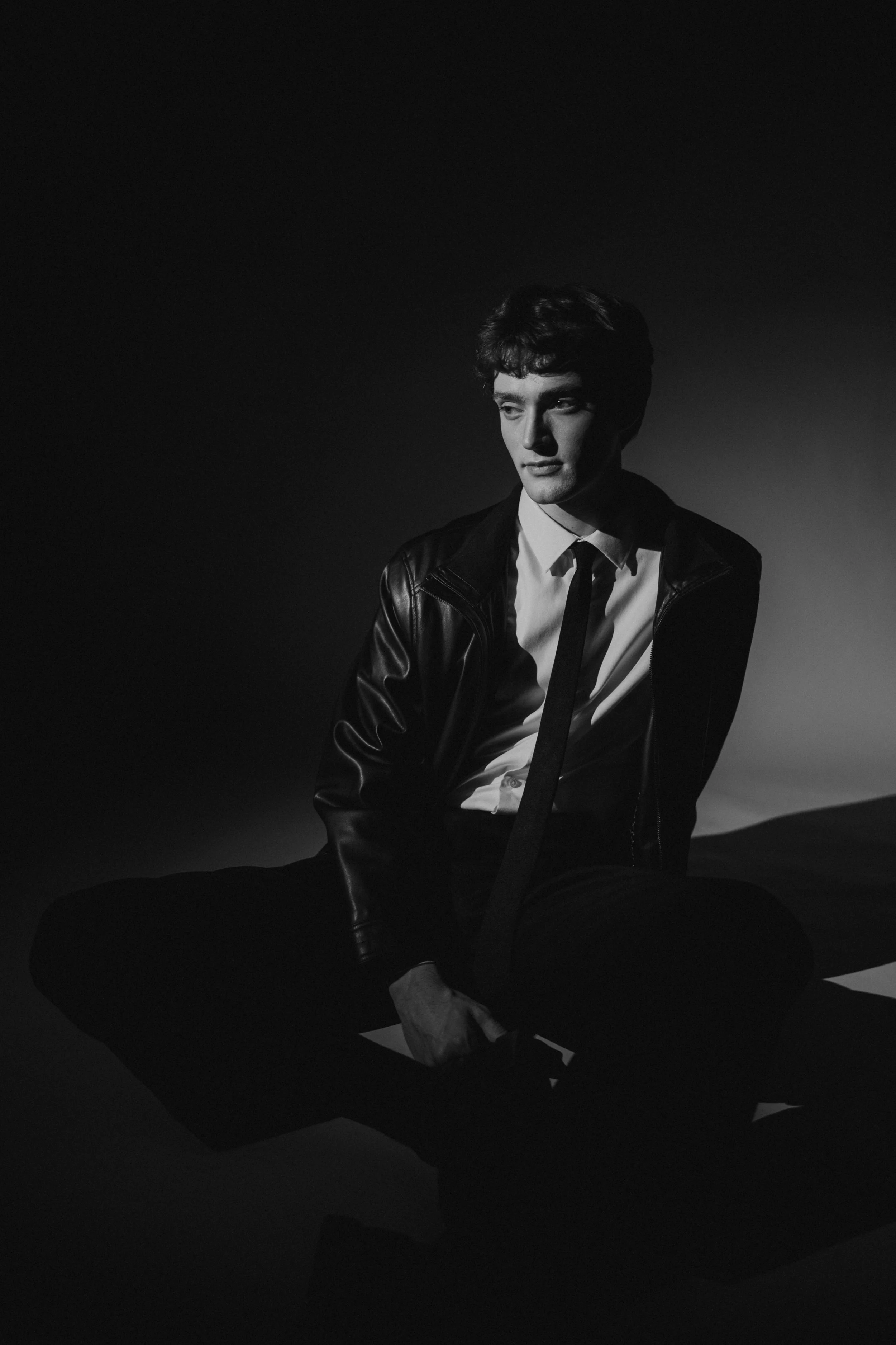 Black and white portrait of a young man sitting on the floor, wearing a leather jacket, white shirt, and tie, with shadows creating a dramatic effect
