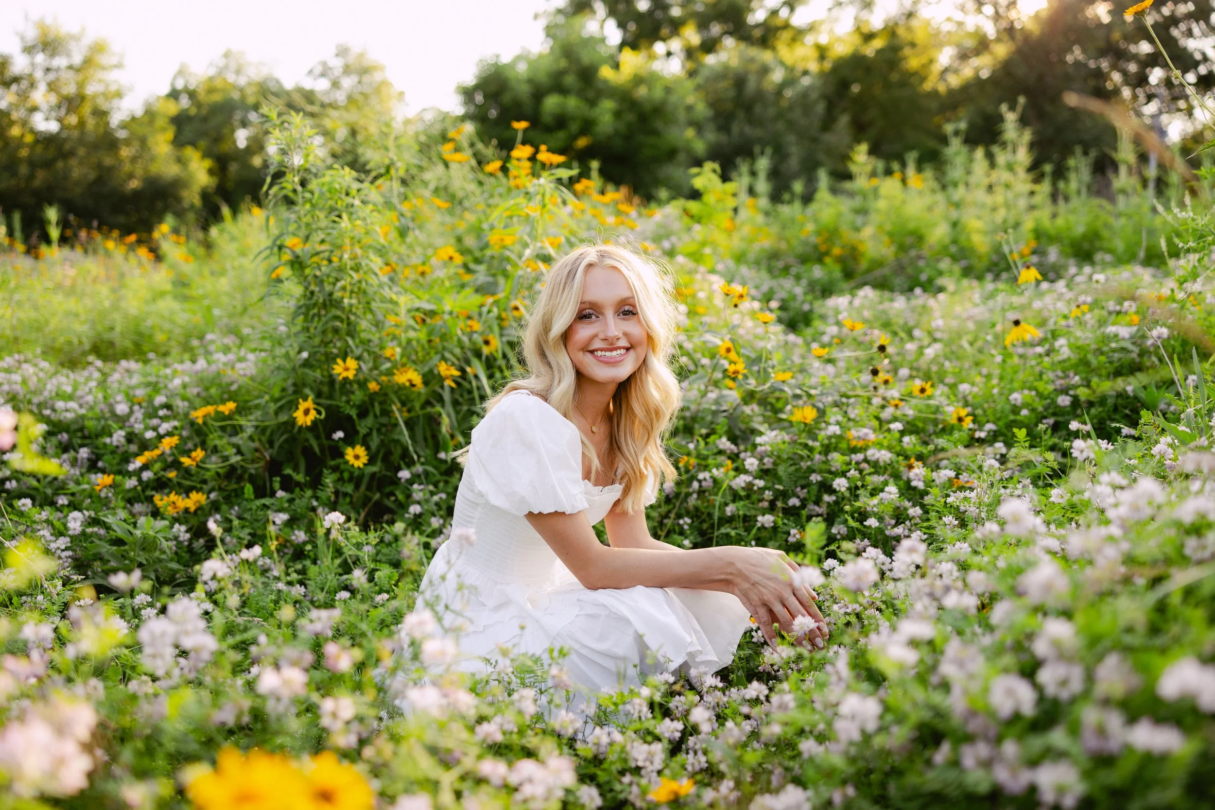 A woman in a white dress sitting among pink and white flowers in a garden with yellow flowers and greenery