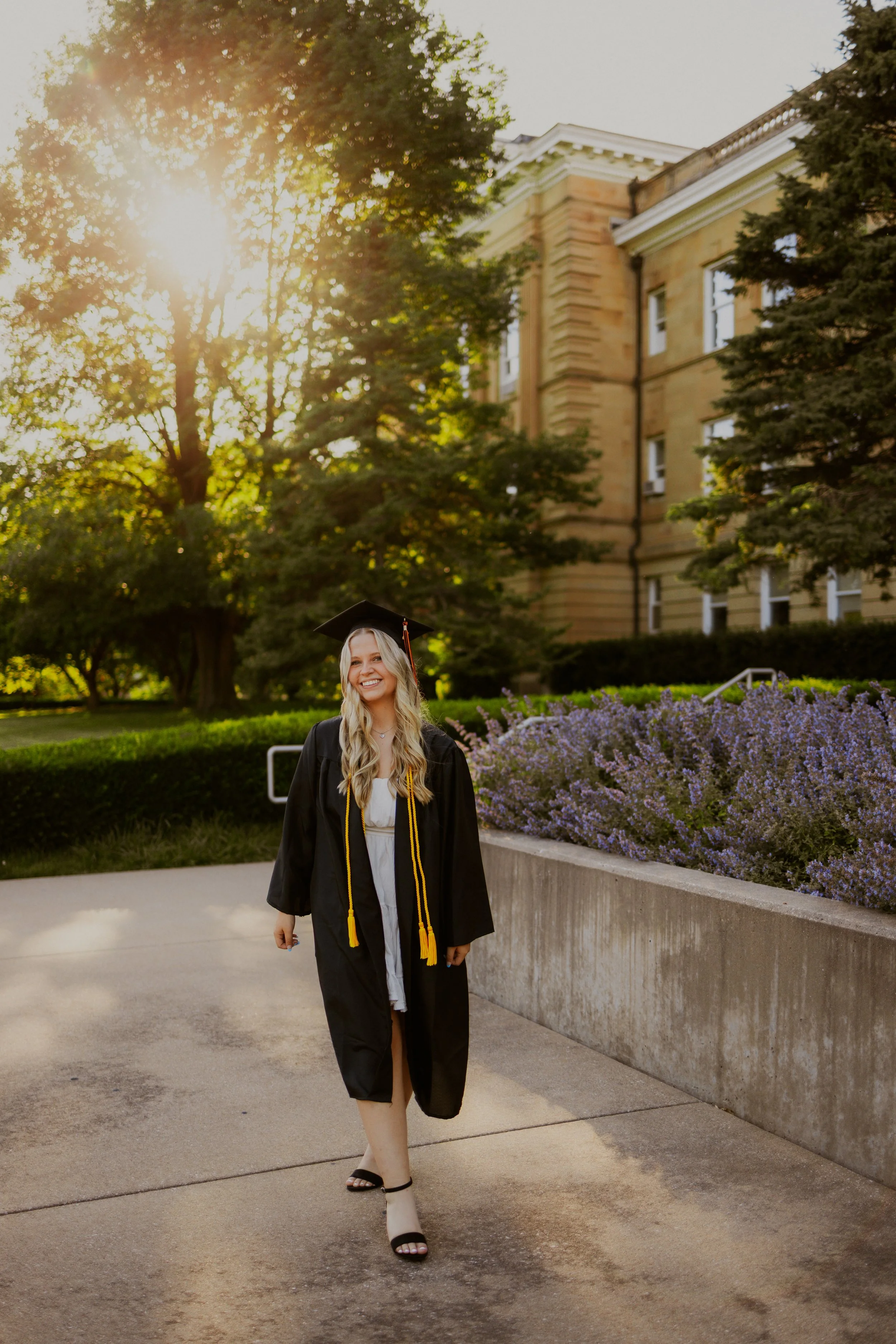 A young woman in a graduation gown and cap, with a yellow honor cord, smiling and walking outdoors on a path lined with purple flowers and green trees, with a large historic building in the background and sunlight filtering through the trees.