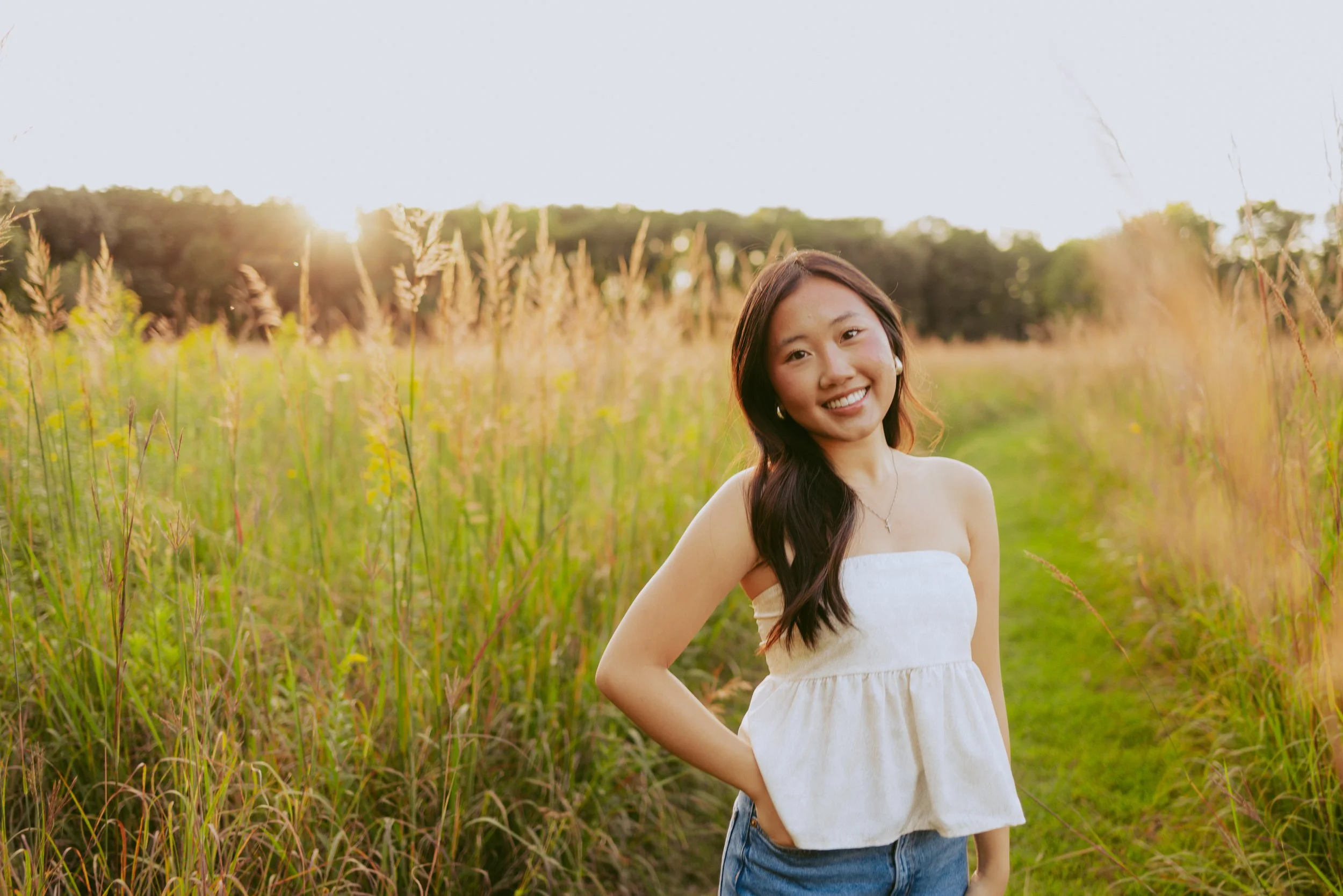 A young woman standing in a grassy field during sunset, smiling at the camera with long dark hair and wearing a white strapless top and blue jeans.