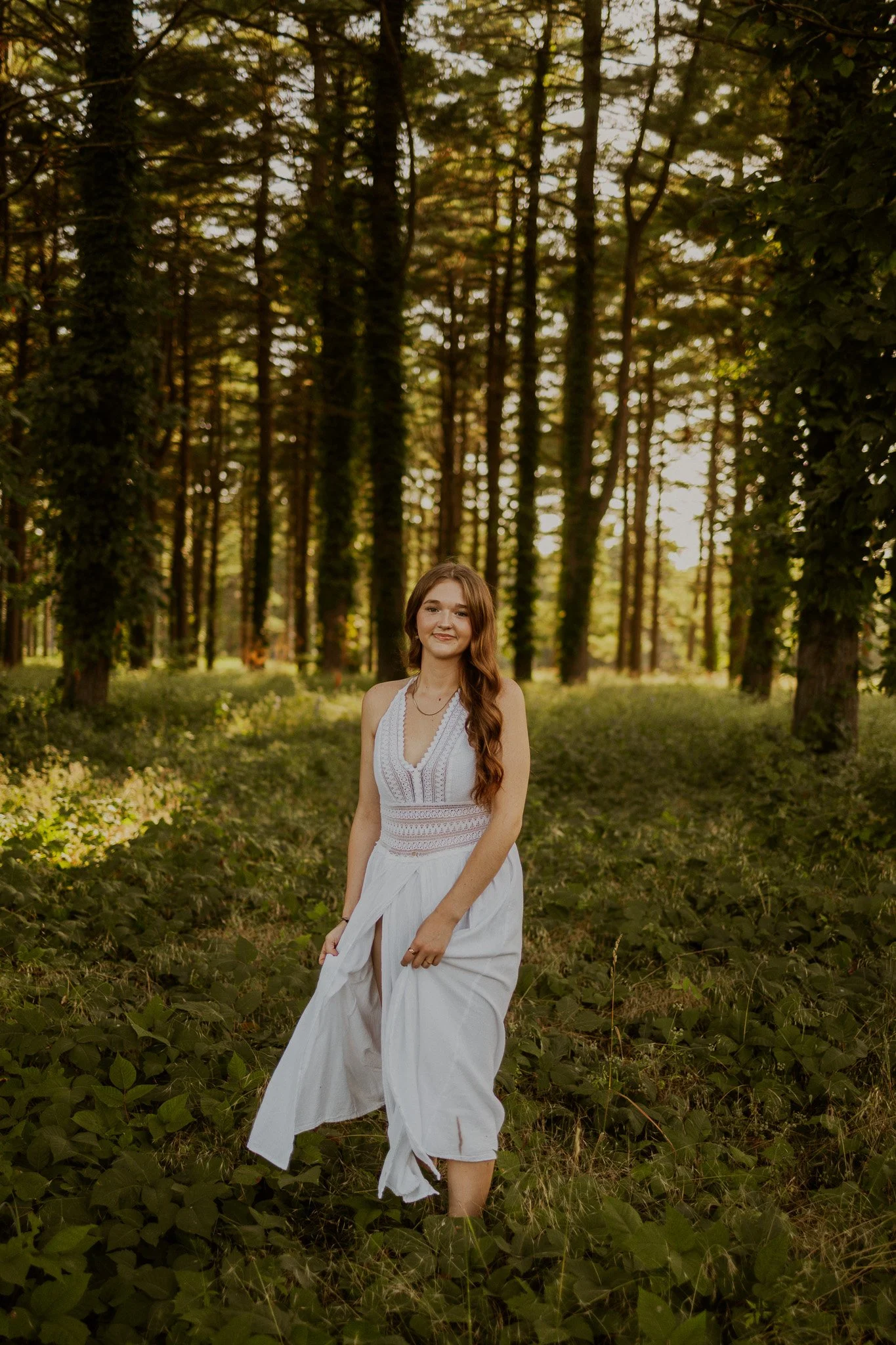 A young woman in a white dress standing in a forest during daylight.
