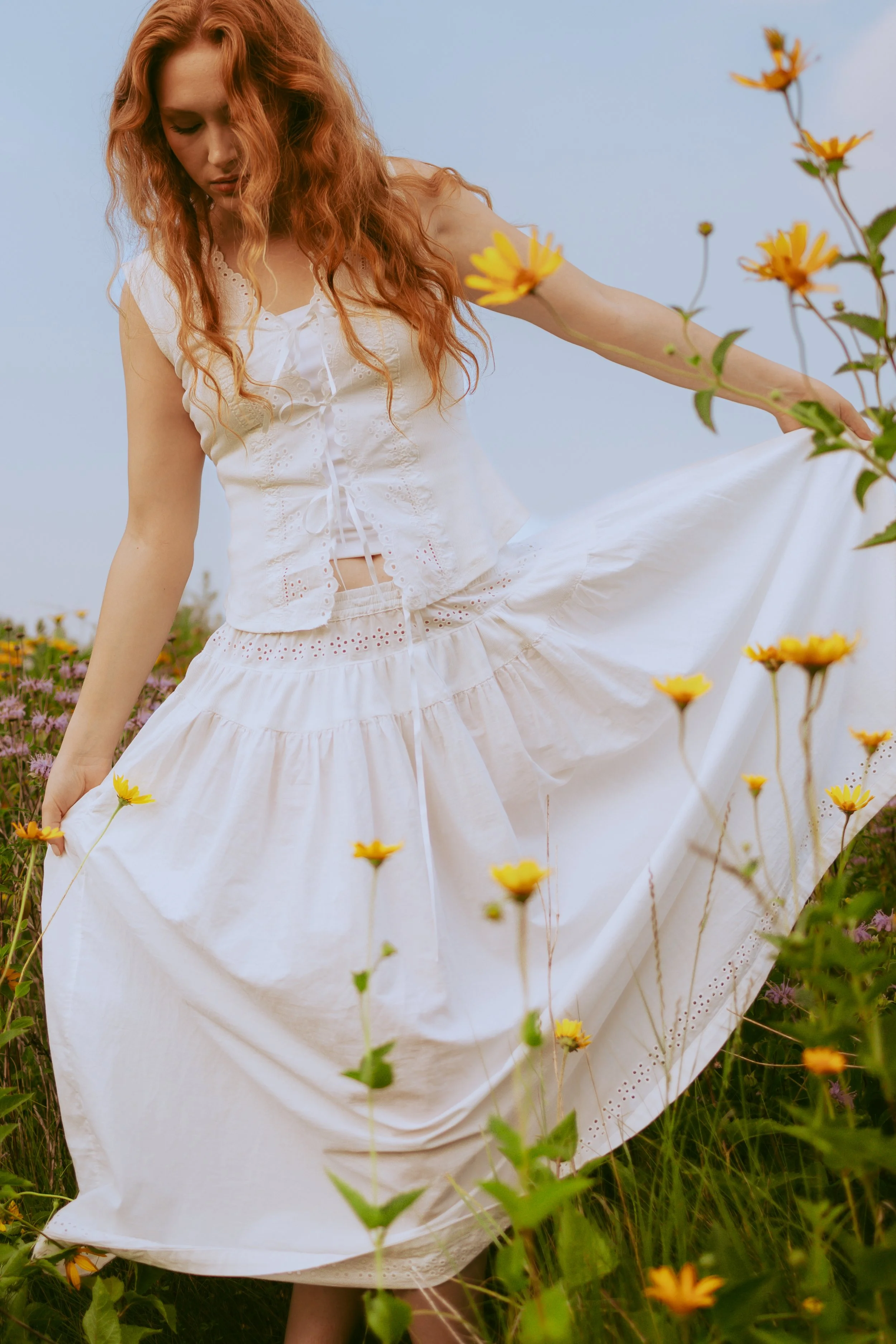 A woman with long, curly red hair stands in a field of yellow and purple flowers, wearing a white dress and looking down.