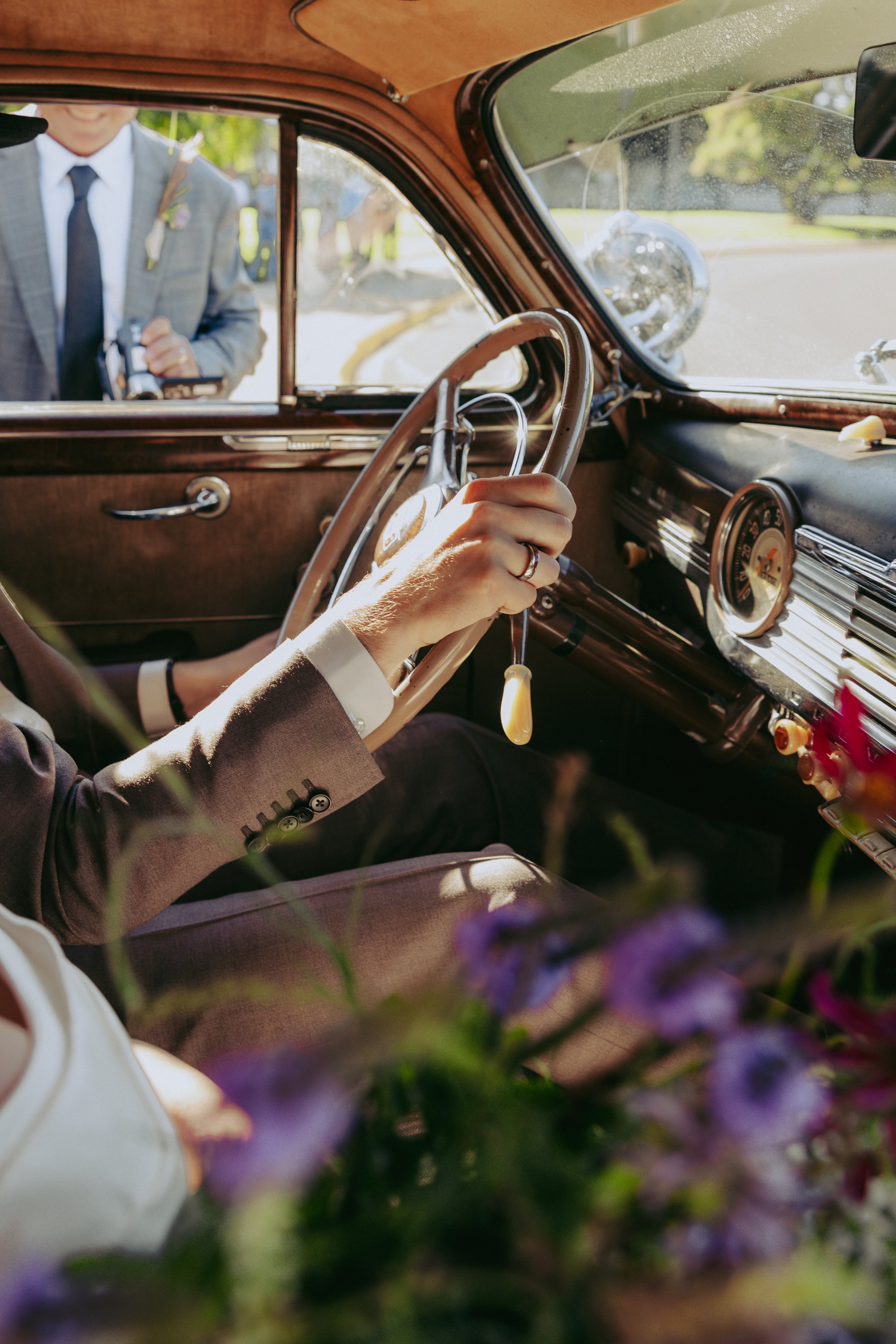 The interior of a vintage car with a person holding the steering wheel, dressed in a suit, with a person outside taking a picture. There are purple flowers in the foreground.