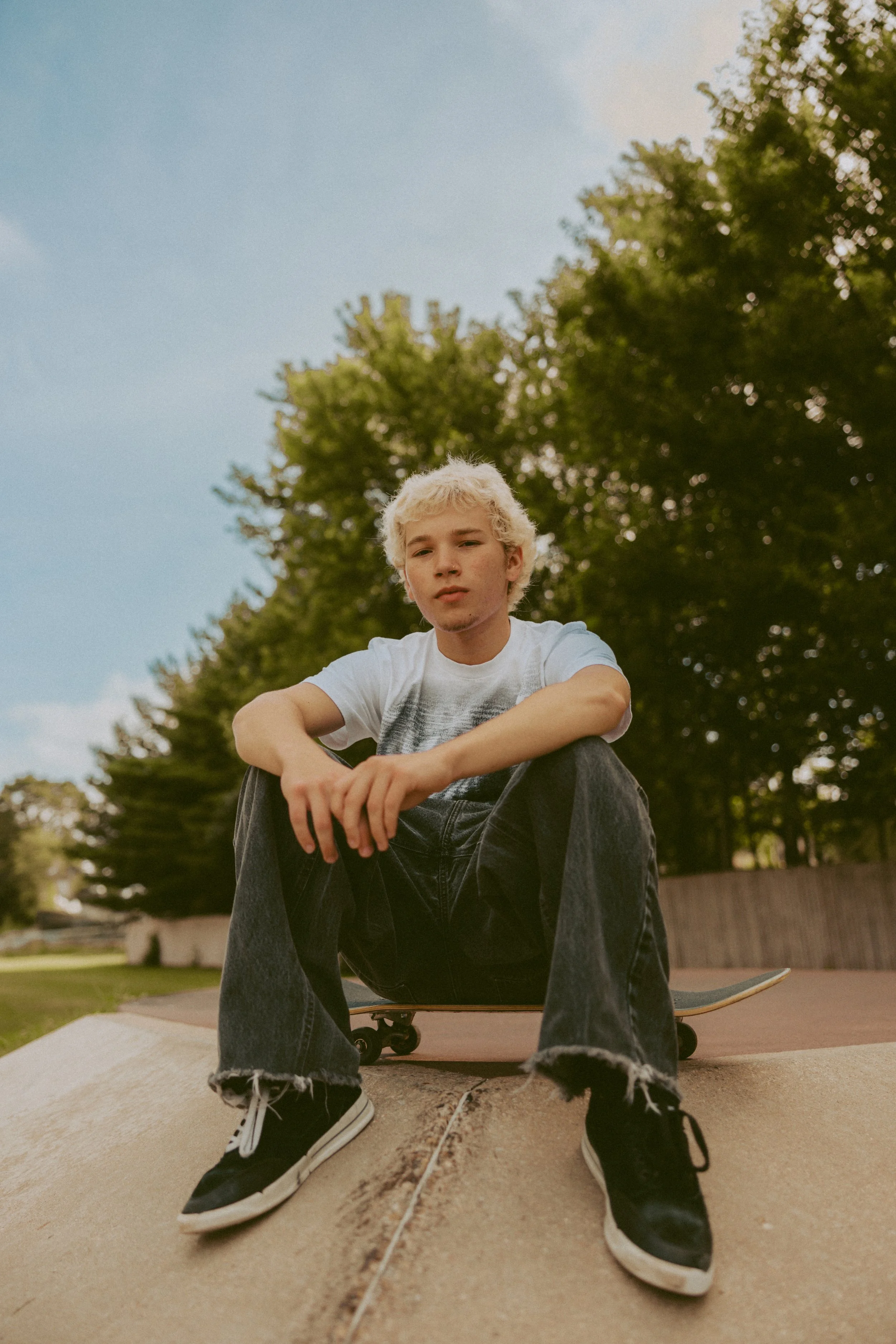 A young male skateboarder with bleach-blonde hair, wearing a white T-shirt, black jeans with frayed hems, and black sneakers, sitting on a skateboard at a skatepark during daytime with clear skies and trees in the background.
