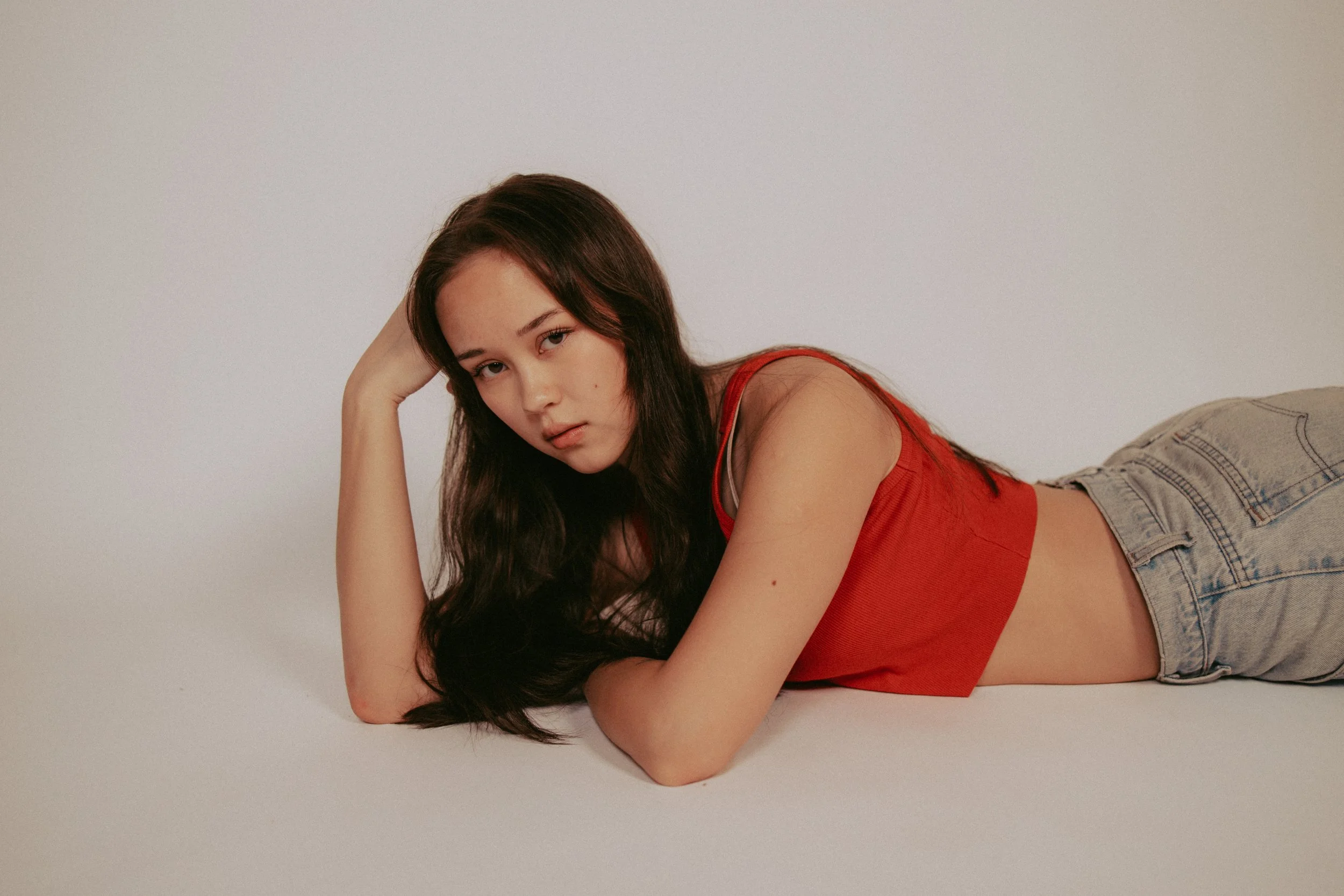 A young woman with long dark hair lying on her side on a plain white surface, wearing a red tank top and light denim jeans, looking directly at the camera.