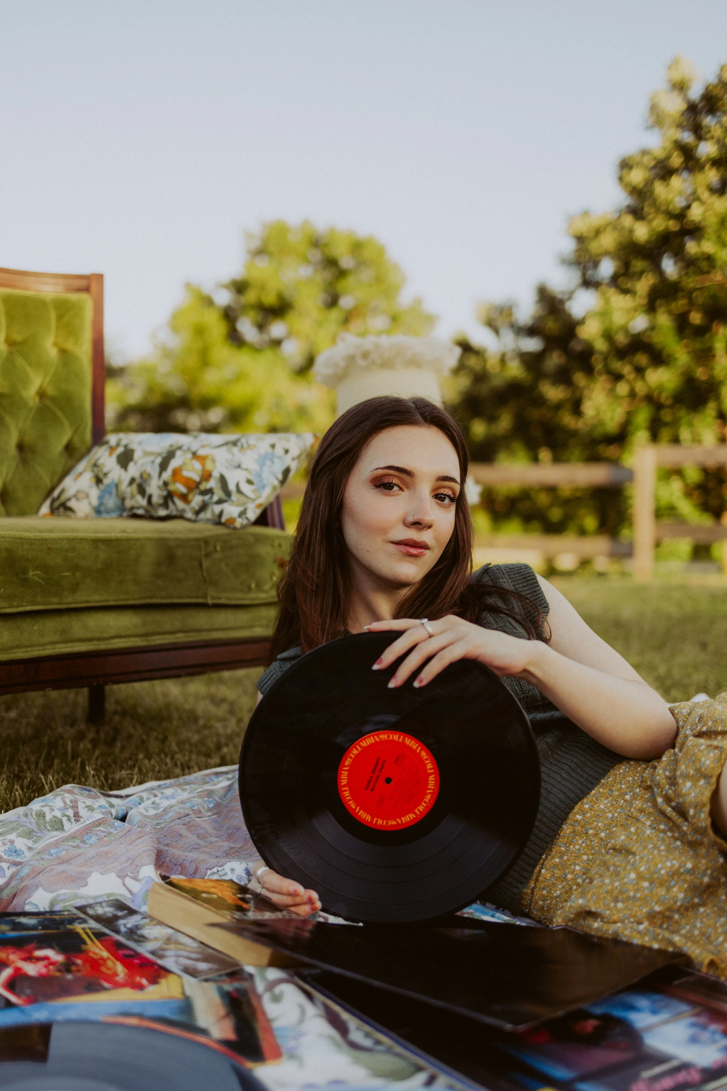 A young woman with brown hair and fair skin lies on the grass outdoors, holding a black vinyl record with a red label. She is near a vintage green velvet couch with floral pillows and a cake on top. The background shows trees and a wooden fence on a 