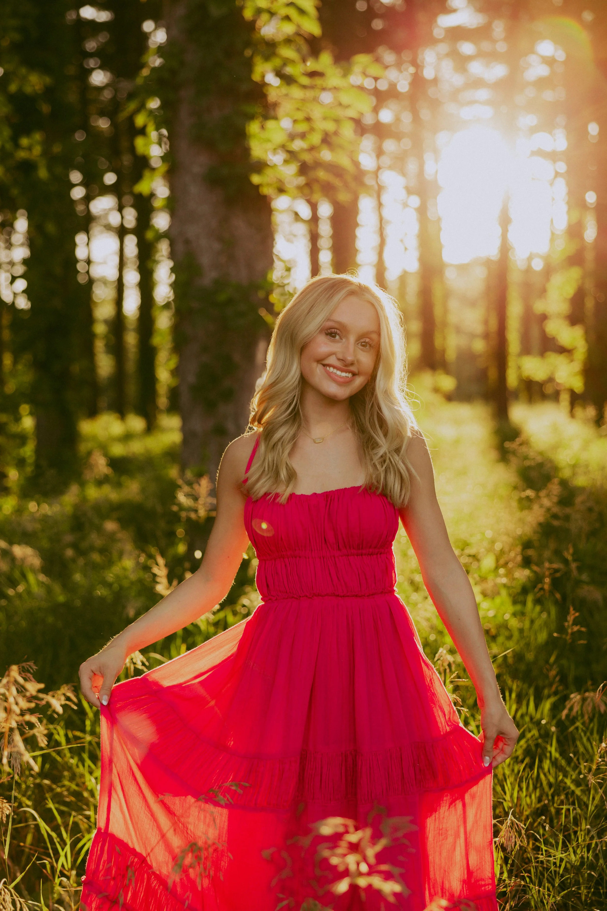 A young woman in a bright pink dress standing in a sunlit forest, smiling and holding the hem of her dress.