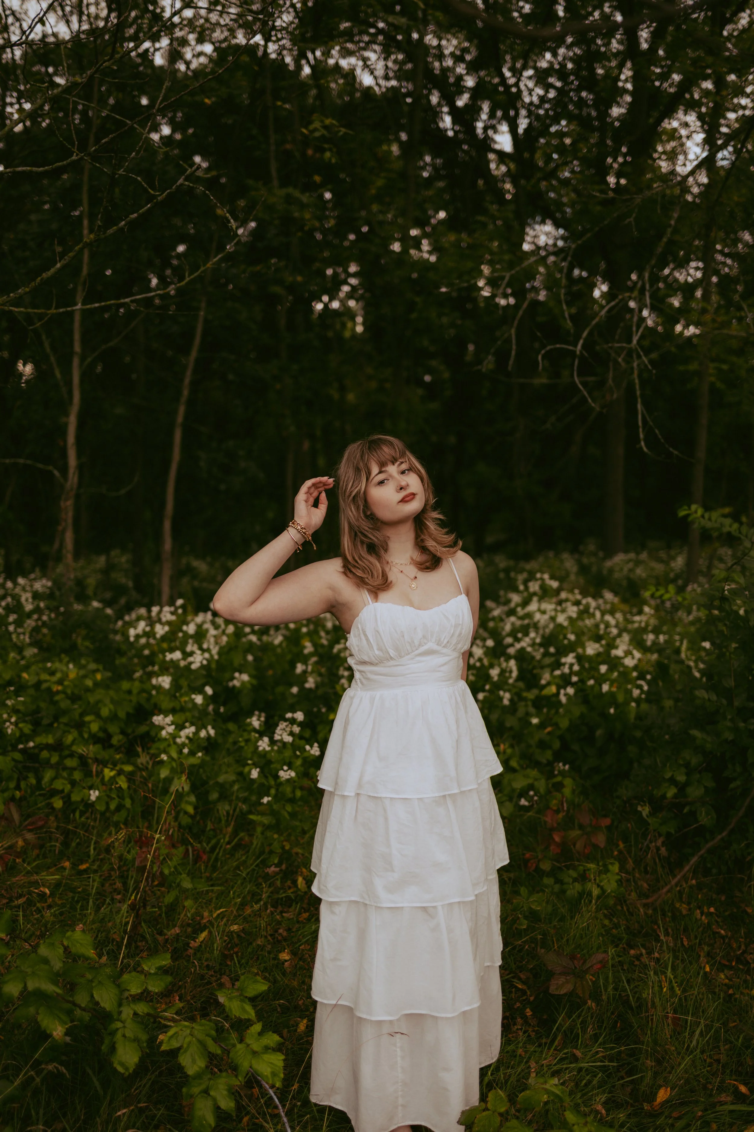 A woman in a white layered dress stands in a forest with green foliage and white flowers, posing with one hand near her head.
