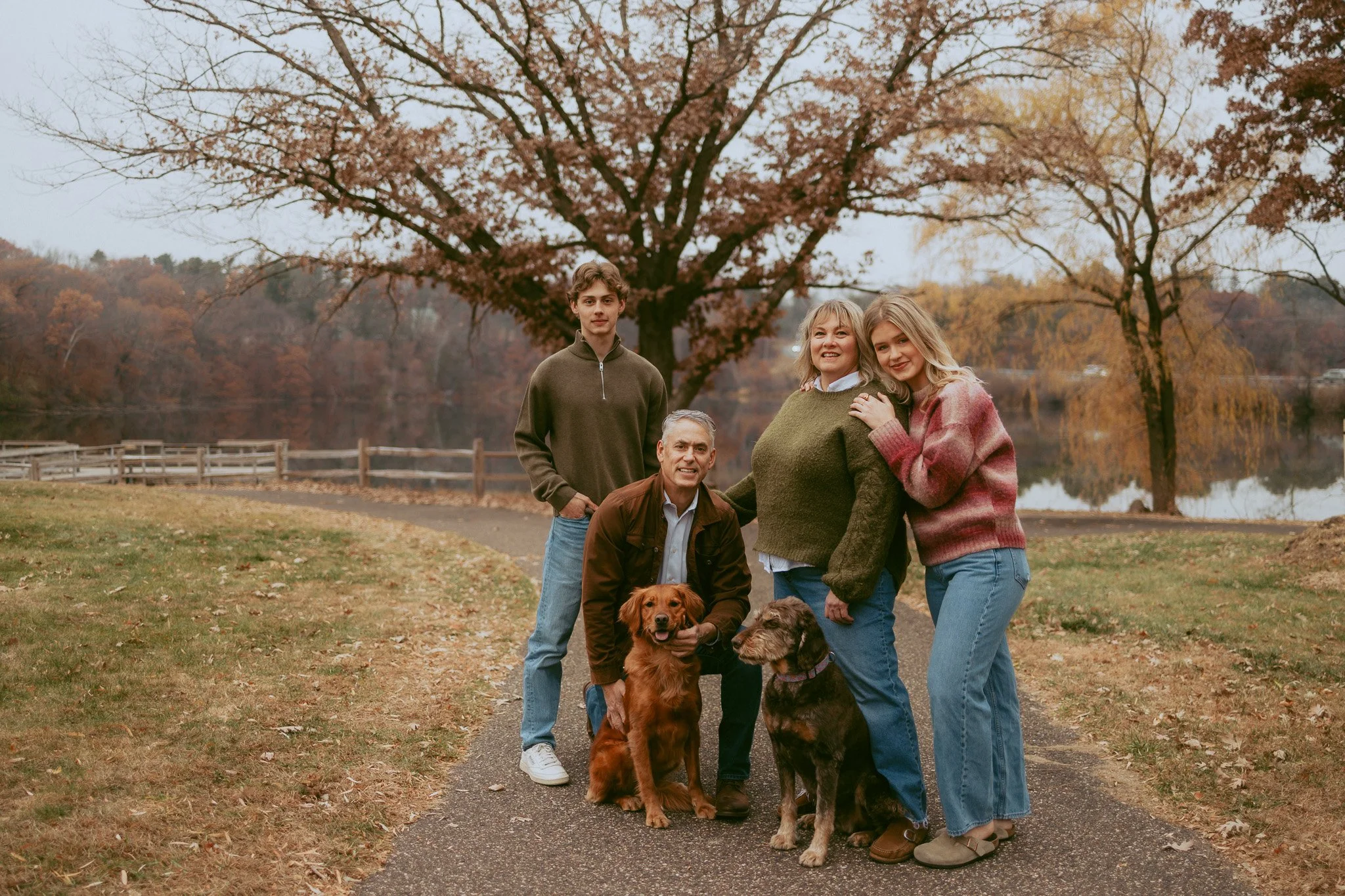 A family of five, including two dogs, standing on a park path with autumn foliage and a lake in the background.
