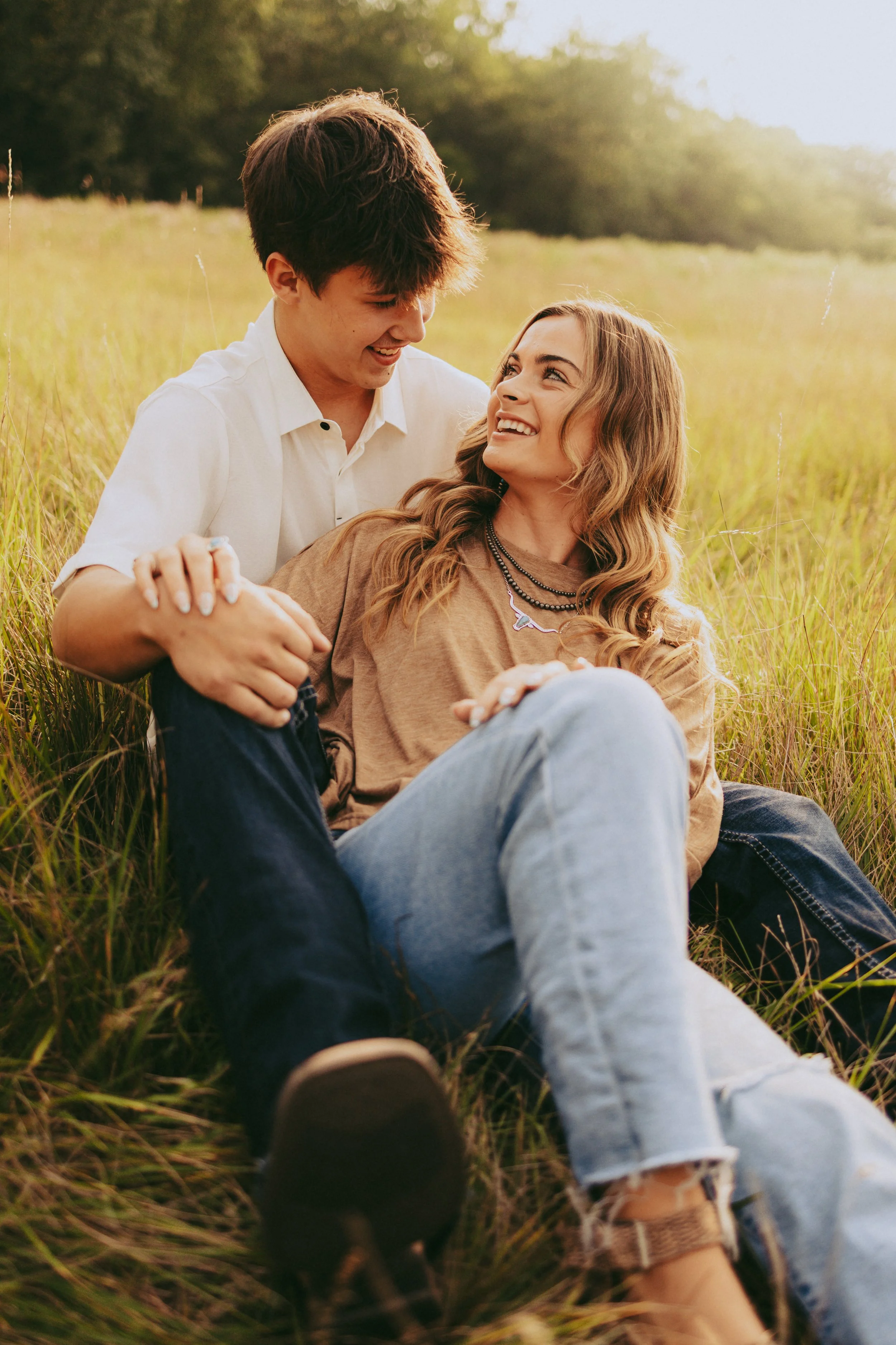 A young couple sitting in a grassy field, smiling and looking at each other during sunset.