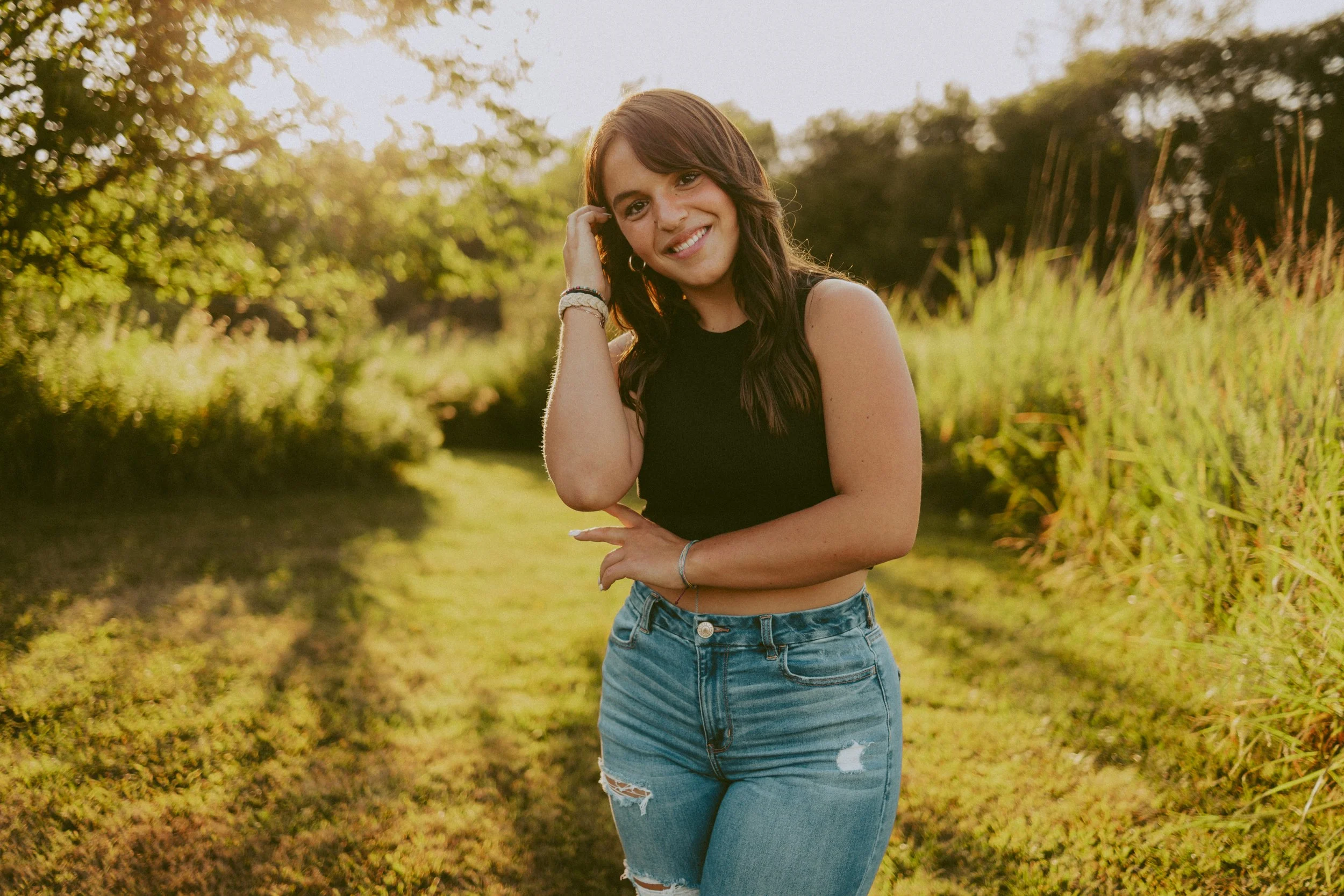 A young woman smiles at the camera, standing outdoors on a grassy path surrounded by trees and bushes with sunlight shining through.