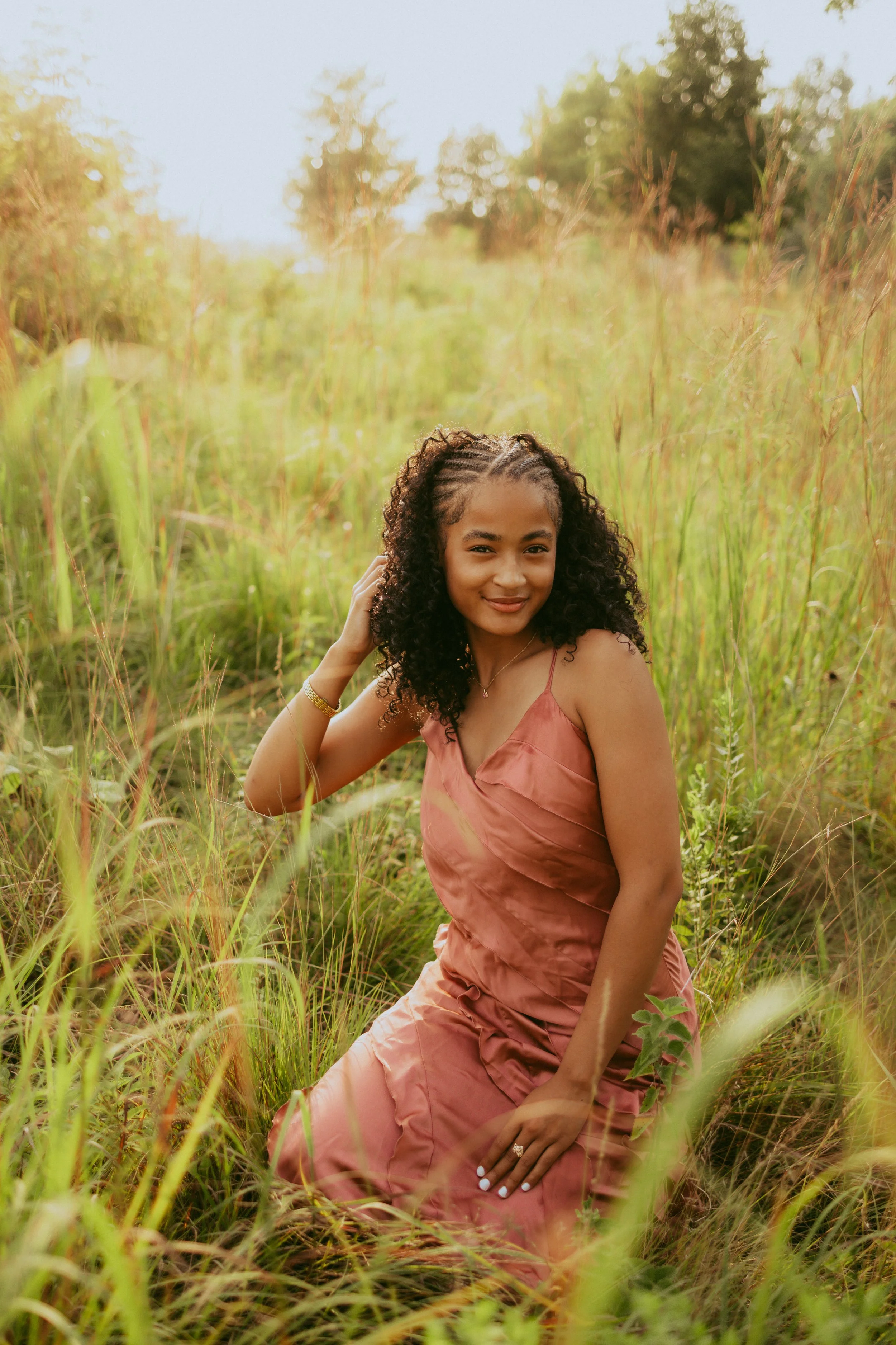 A woman with curly hair in a pink dress kneeling in tall grass in a field during golden hour.
