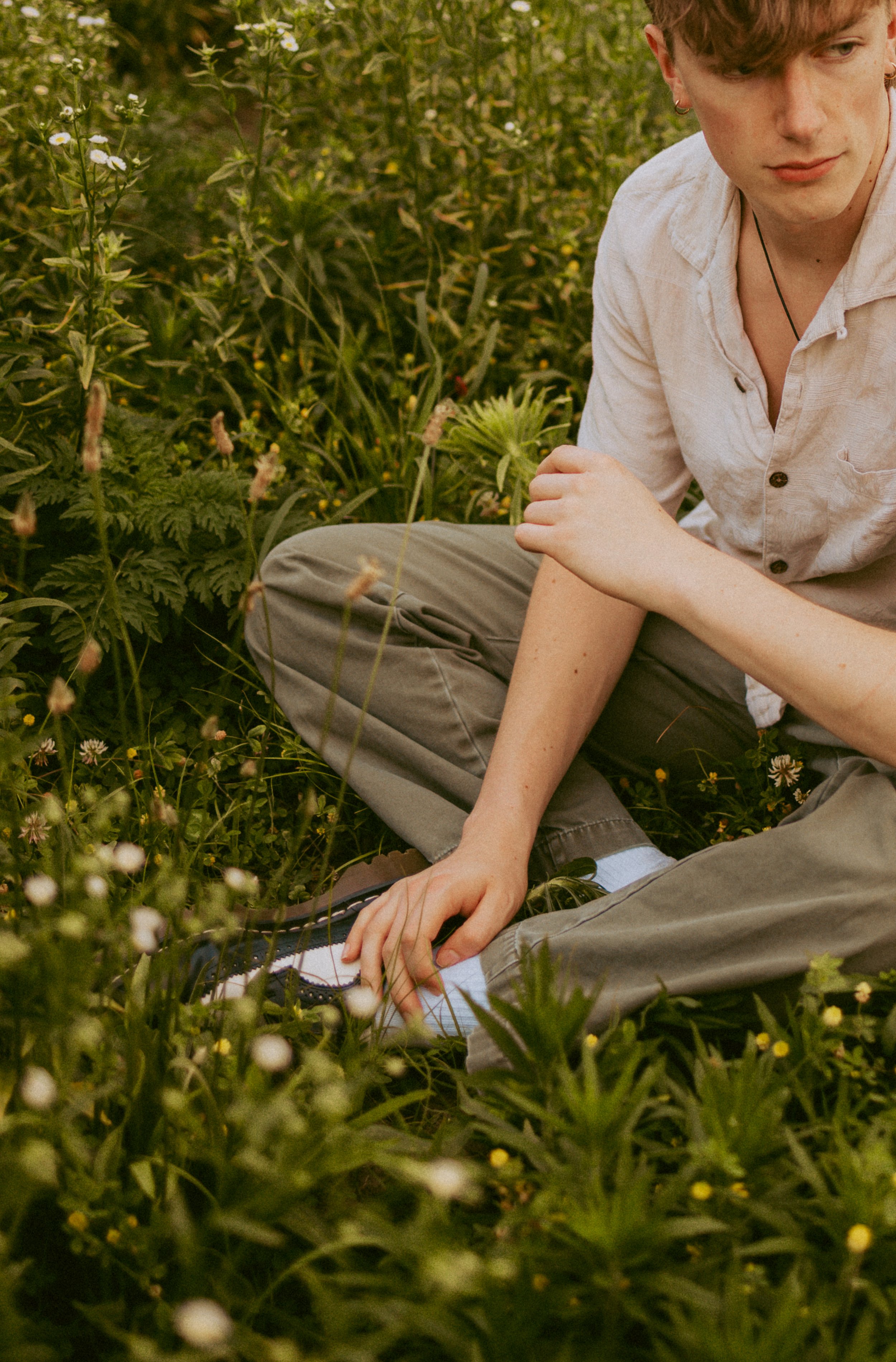 A young man is sitting on the ground among green plants and flowers, wearing a light-colored shirt, brown pants, and hiking shoes.