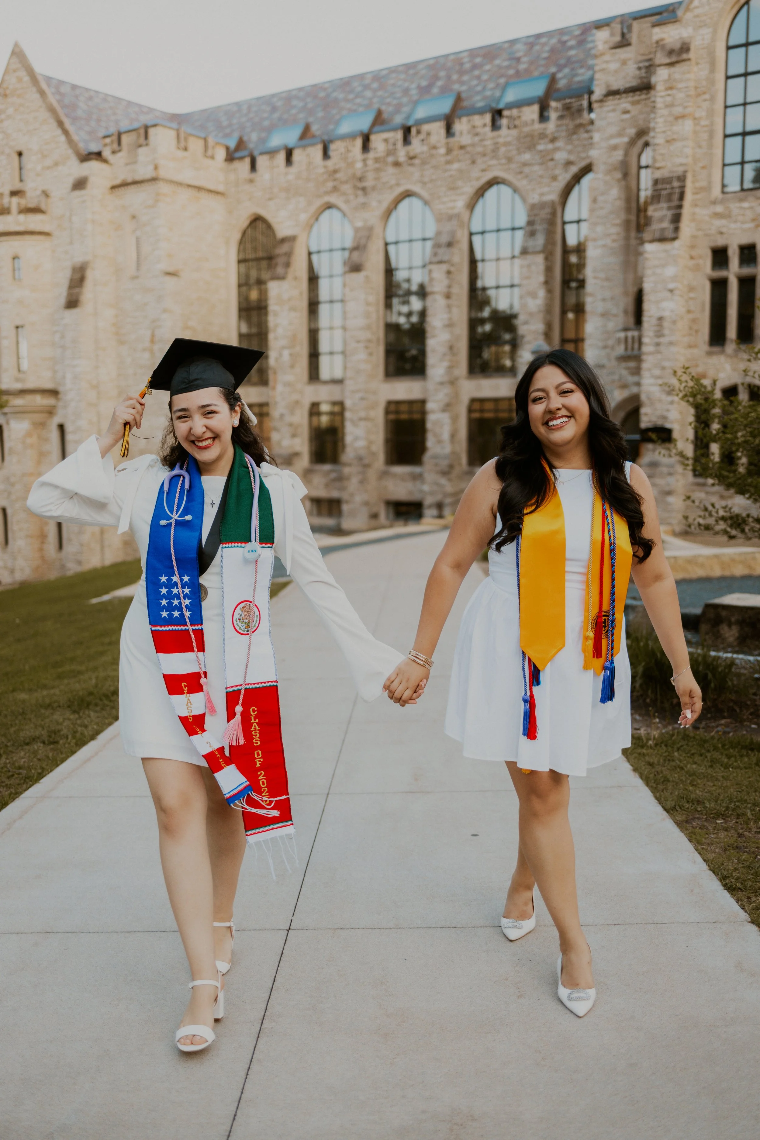 Two women in graduation caps and dresses walking hand in hand outside a campus building, celebrating graduation.