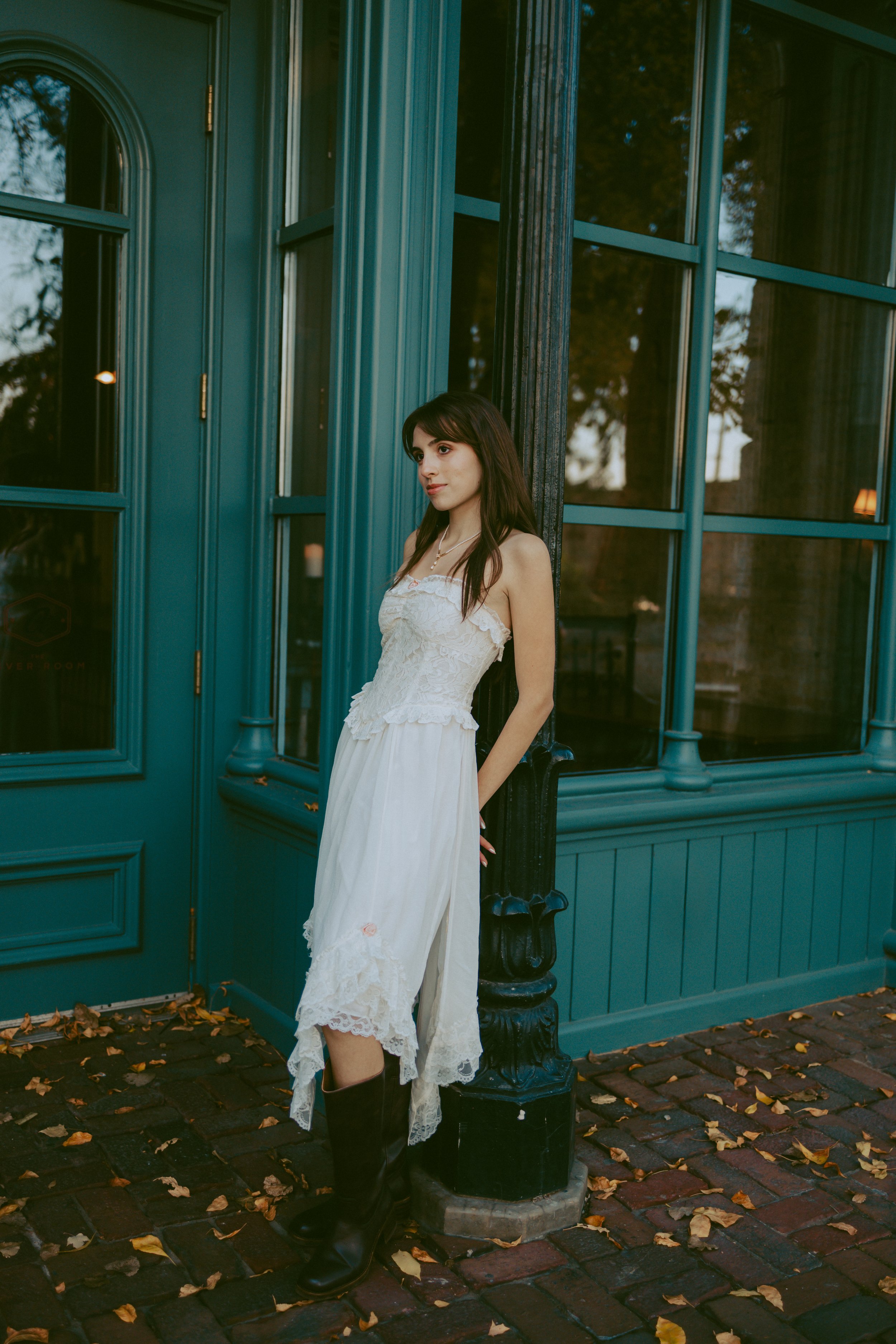 A young woman in a white dress and black boots leaning against a black lamppost outside a teal building with large windows.