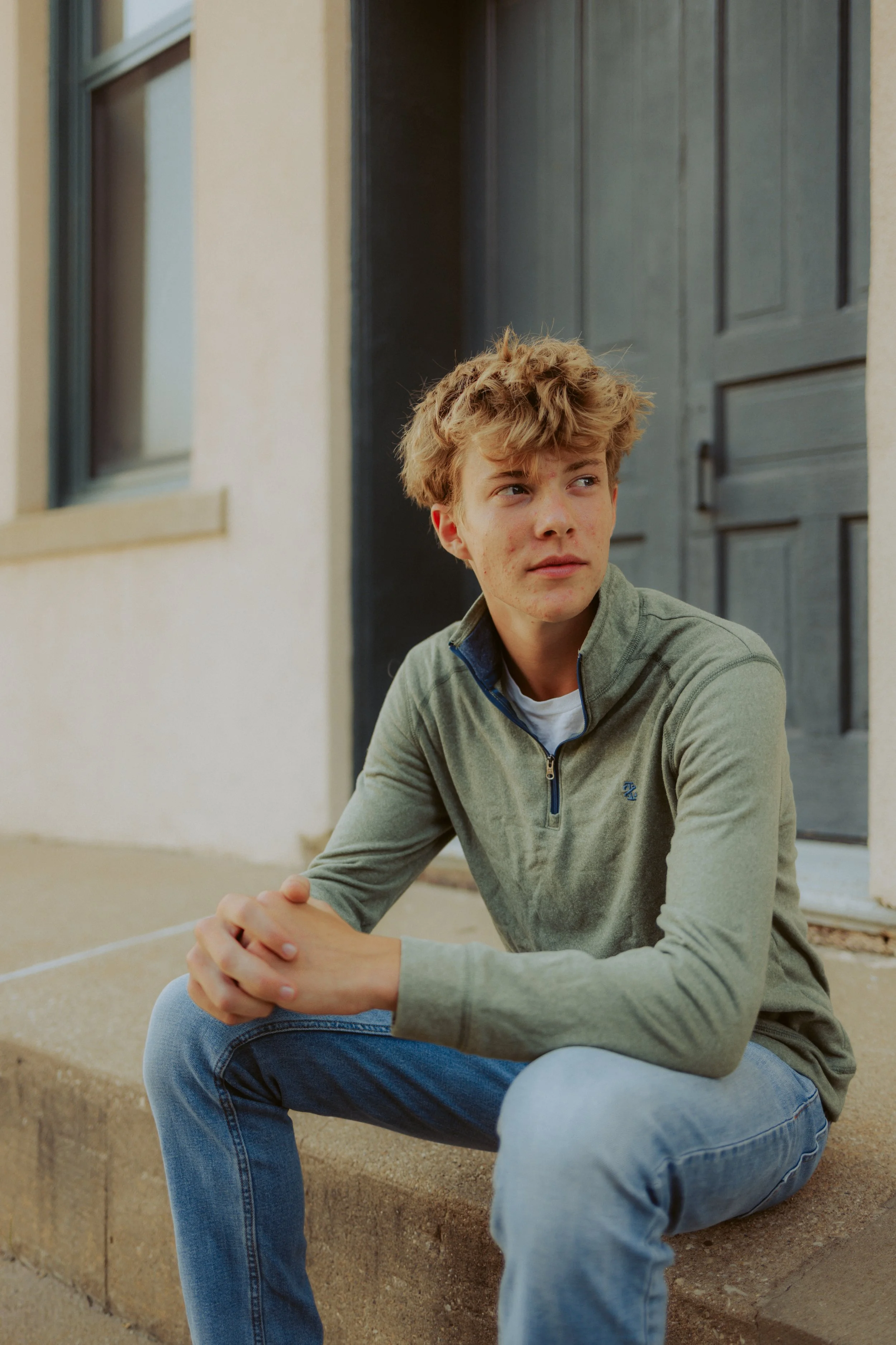 A teenage boy with light brown, tousled hair, wearing a light green quarter-zip sweater and blue jeans, sitting on concrete steps outside a building with a dark gray door, looking to the side with a neutral expression.
