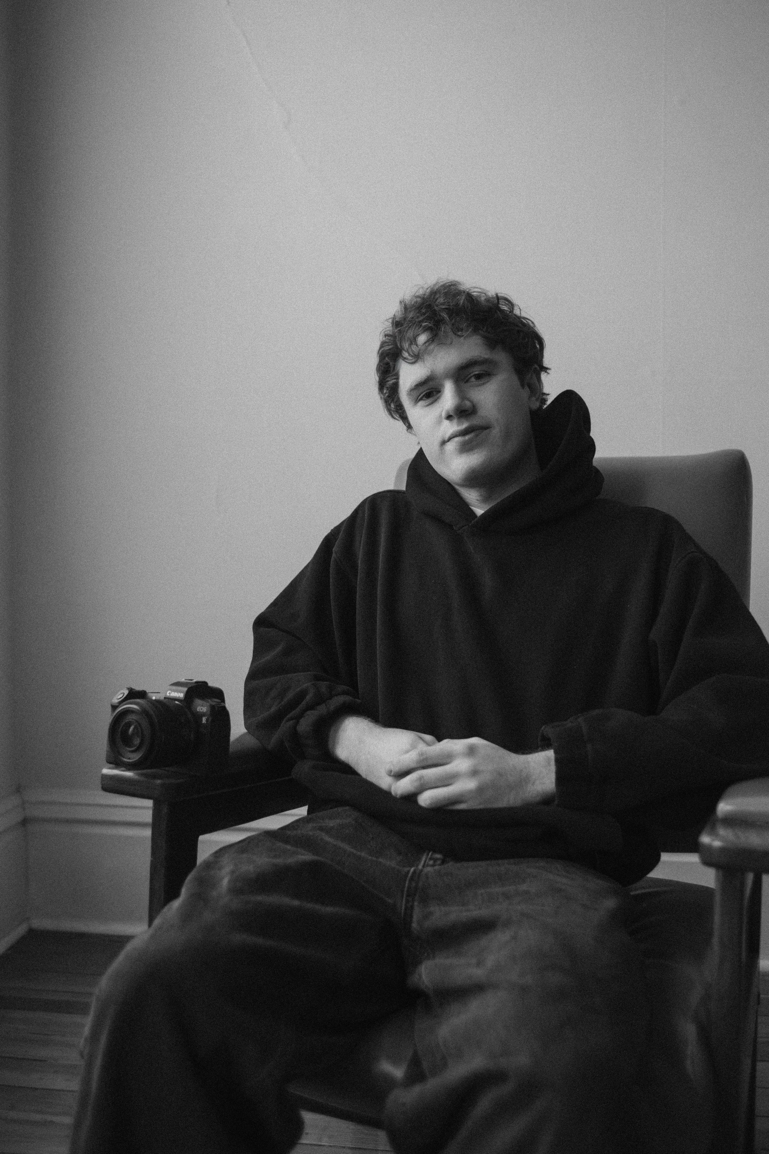 A young man with curly hair wearing a black hoodie, sitting on a chair next to a camera on a small table, in a room with plain walls.