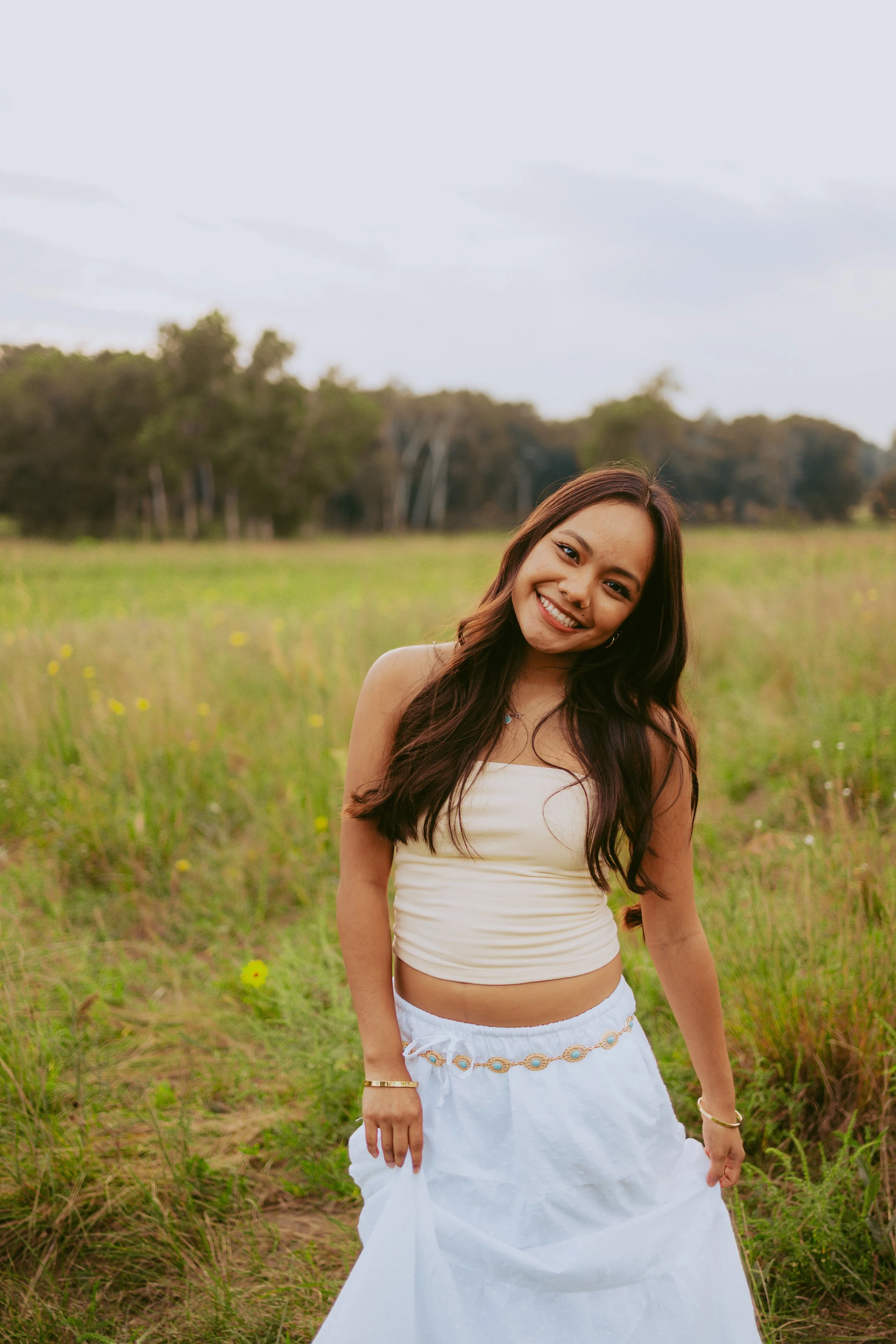 A young woman with long dark hair standing in a green field, smiling at the camera, wearing a white crop top and a long white skirt.