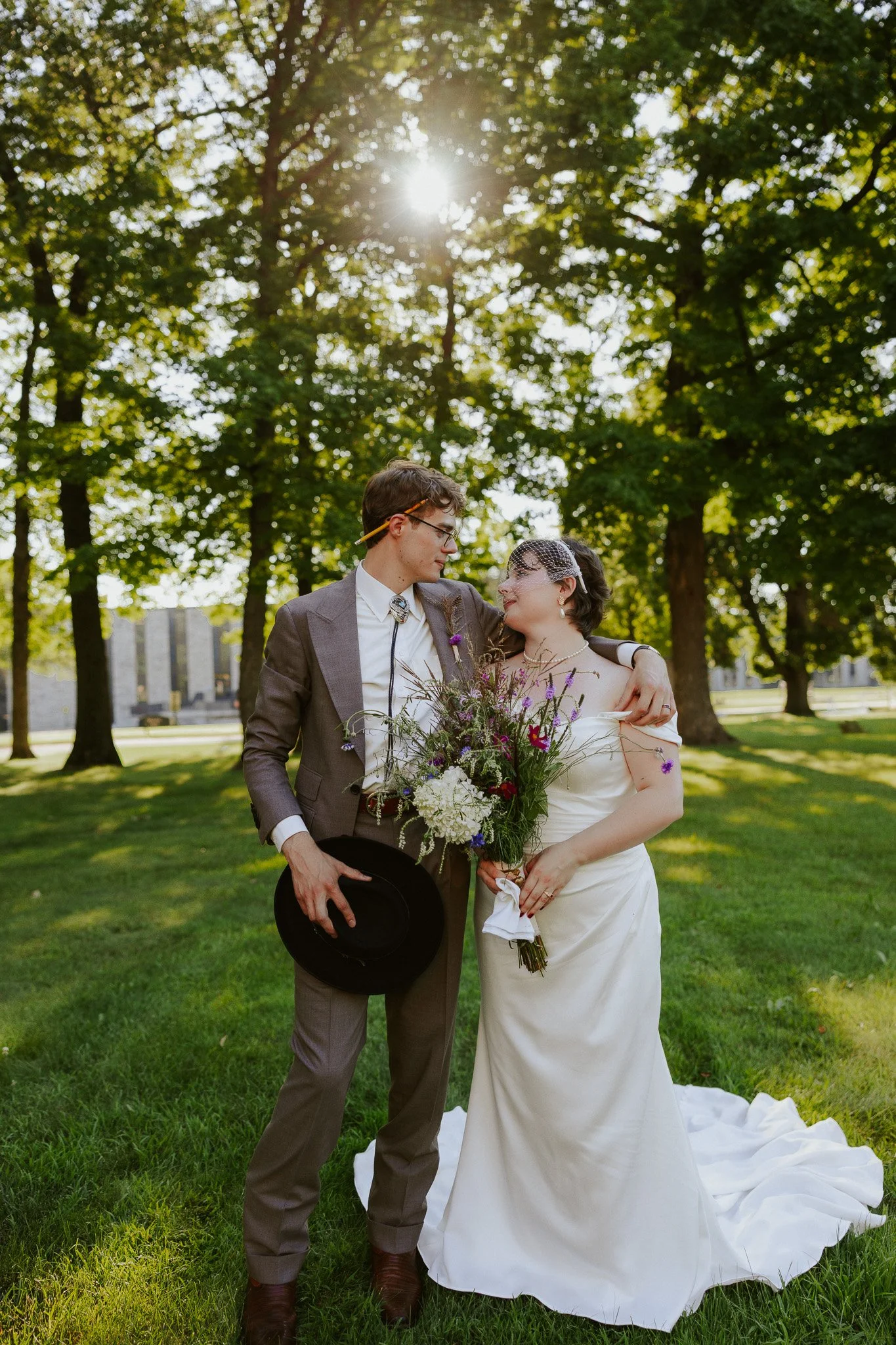 A wedding couple in a park with trees and sunlight, the groom wearing a suit and glasses holding a hat, and the bride in a white dress holding a bouquet of flowers, looking at each other lovingly.