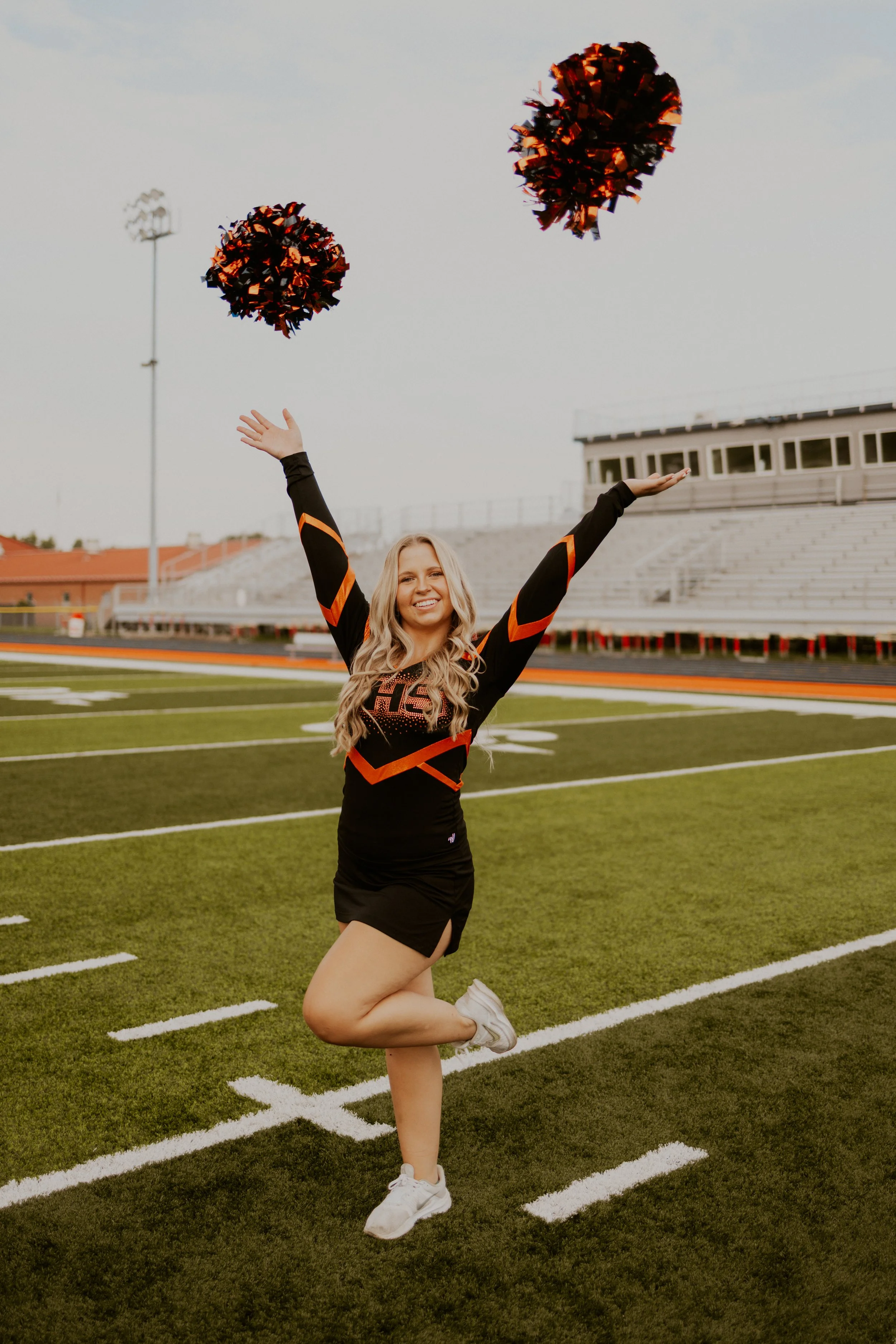 A cheerleader in a black and orange uniform is standing on a football field, smiling, and jumping with one leg raised, throwing black and orange pom-poms into the air.