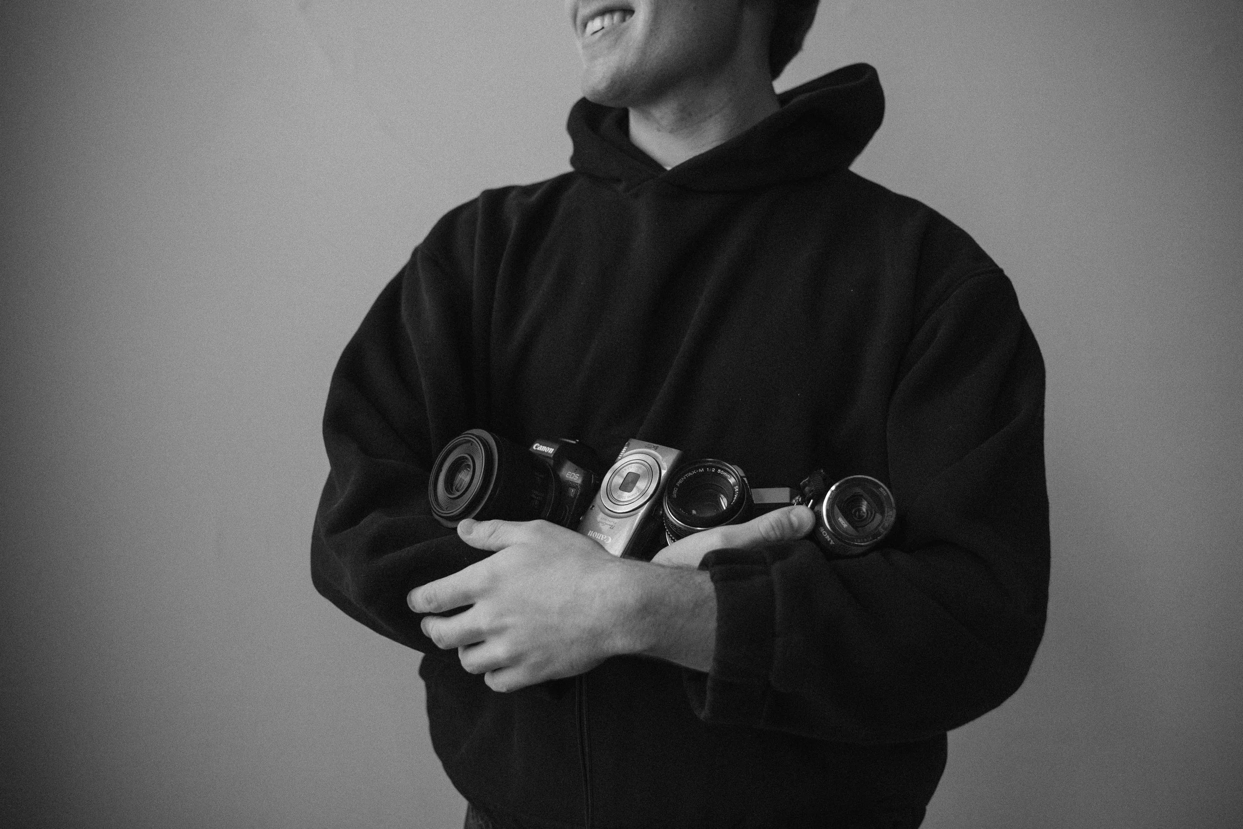 Person in a black hoodie holding three cameras against a plain background.