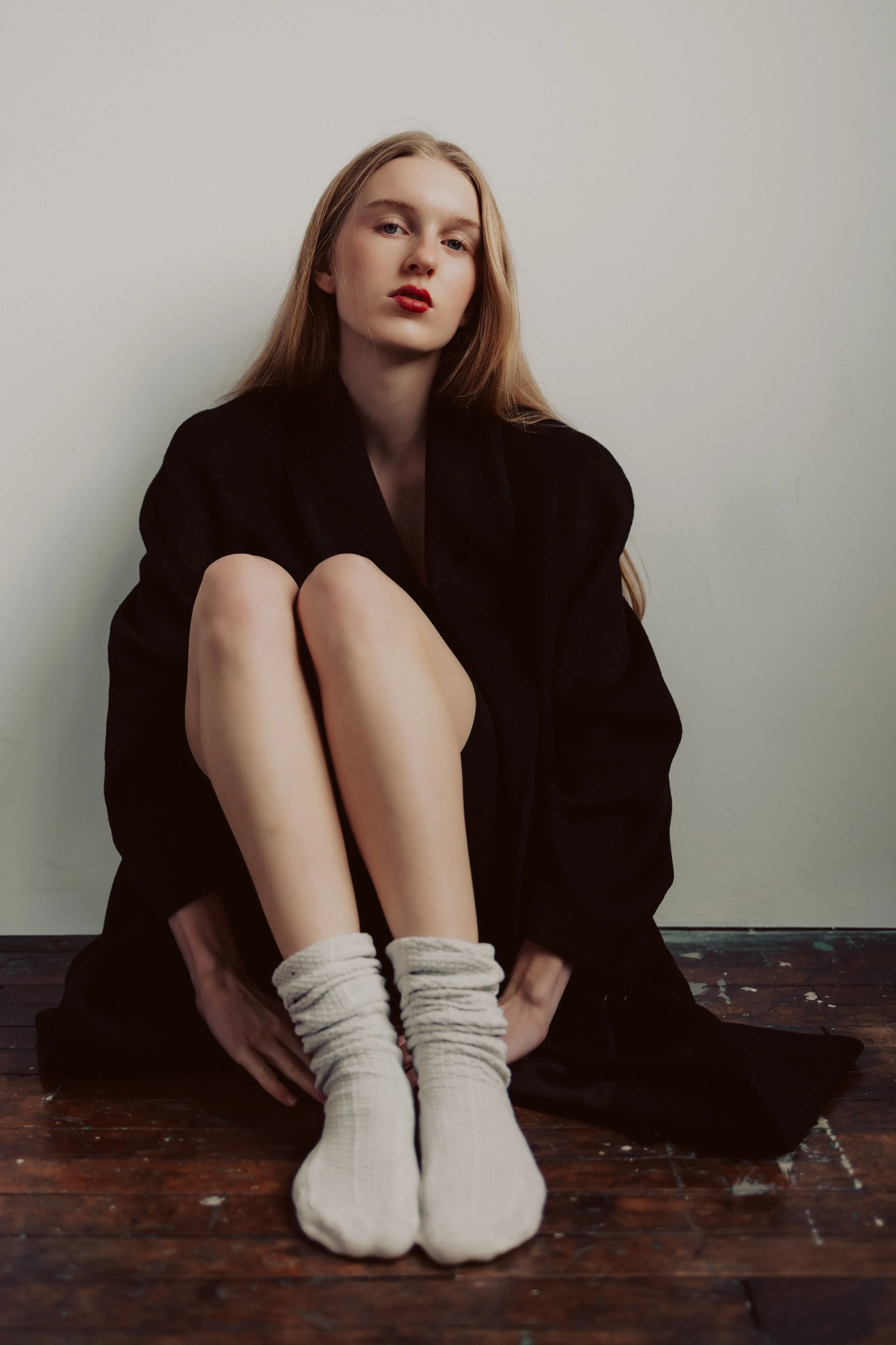 A young woman with long blonde hair, wearing red lipstick, sitting on a wooden floor with her knees pulled up to her chest, dressed in a black coat and white socks, against a plain white wall.