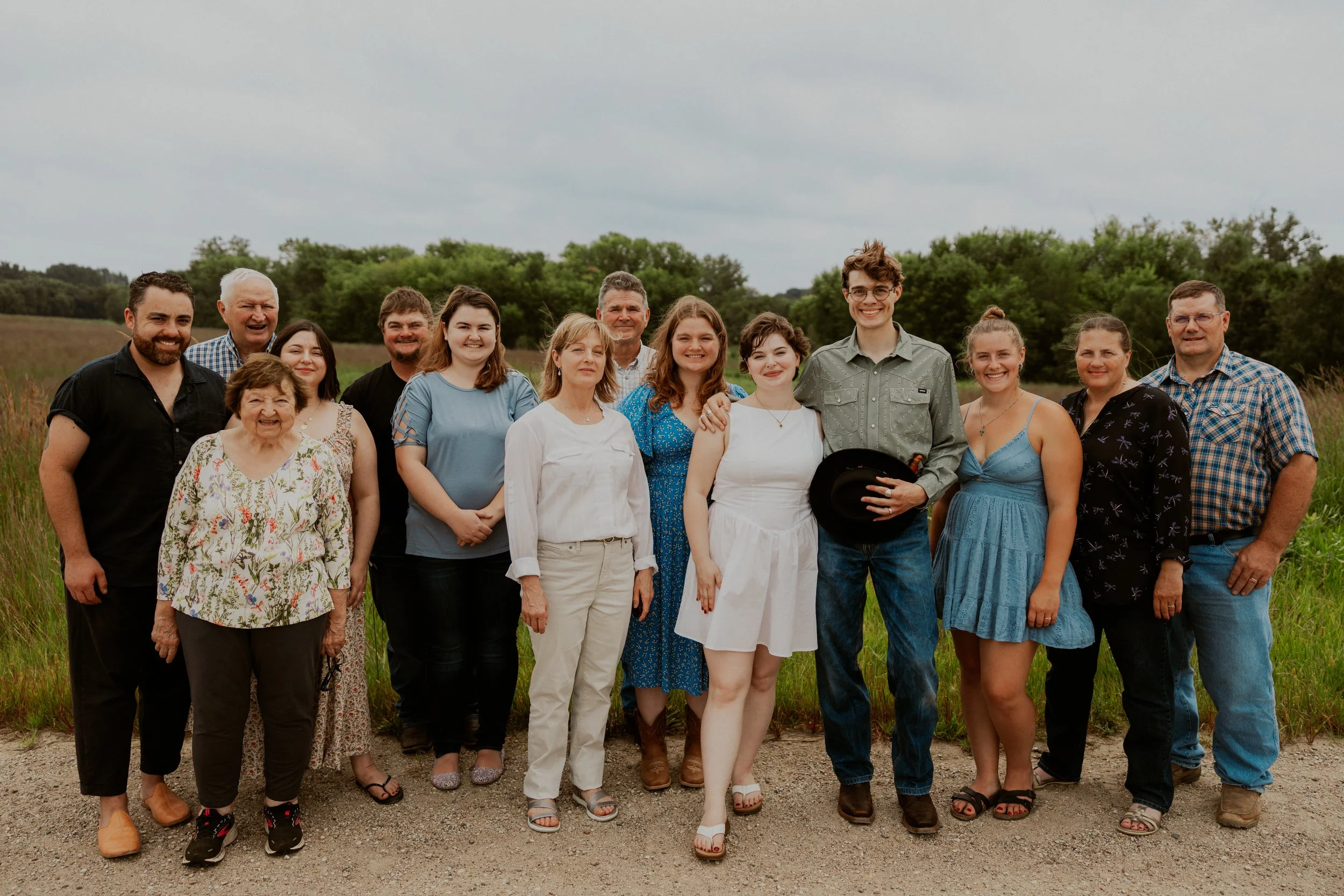 A large group of family members standing outdoors on a gravel road with green fields and trees in the background, smiling for a photo.