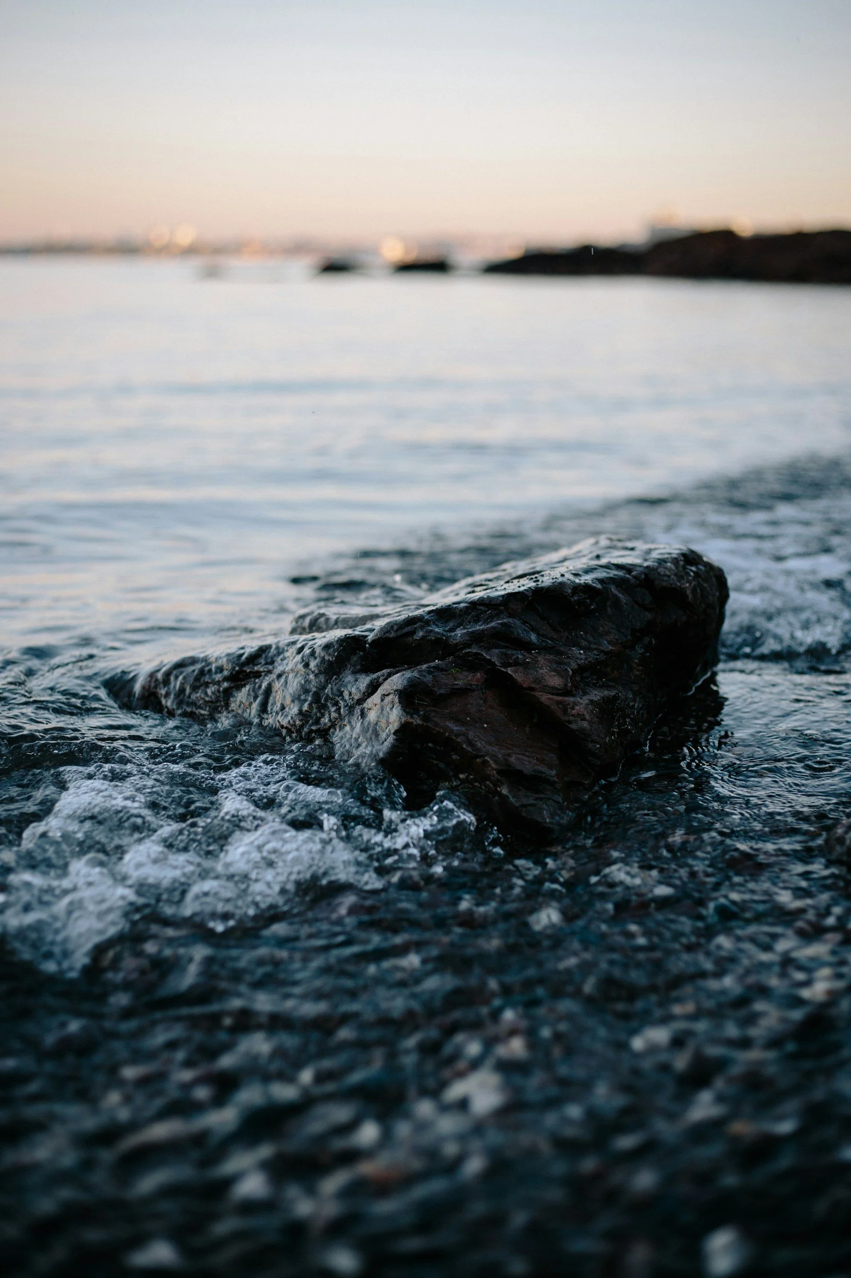 Close-up view of a dark, wet rock in the water near the shoreline, with a blurred background of the ocean and a distant horizon during dusk or dawn.