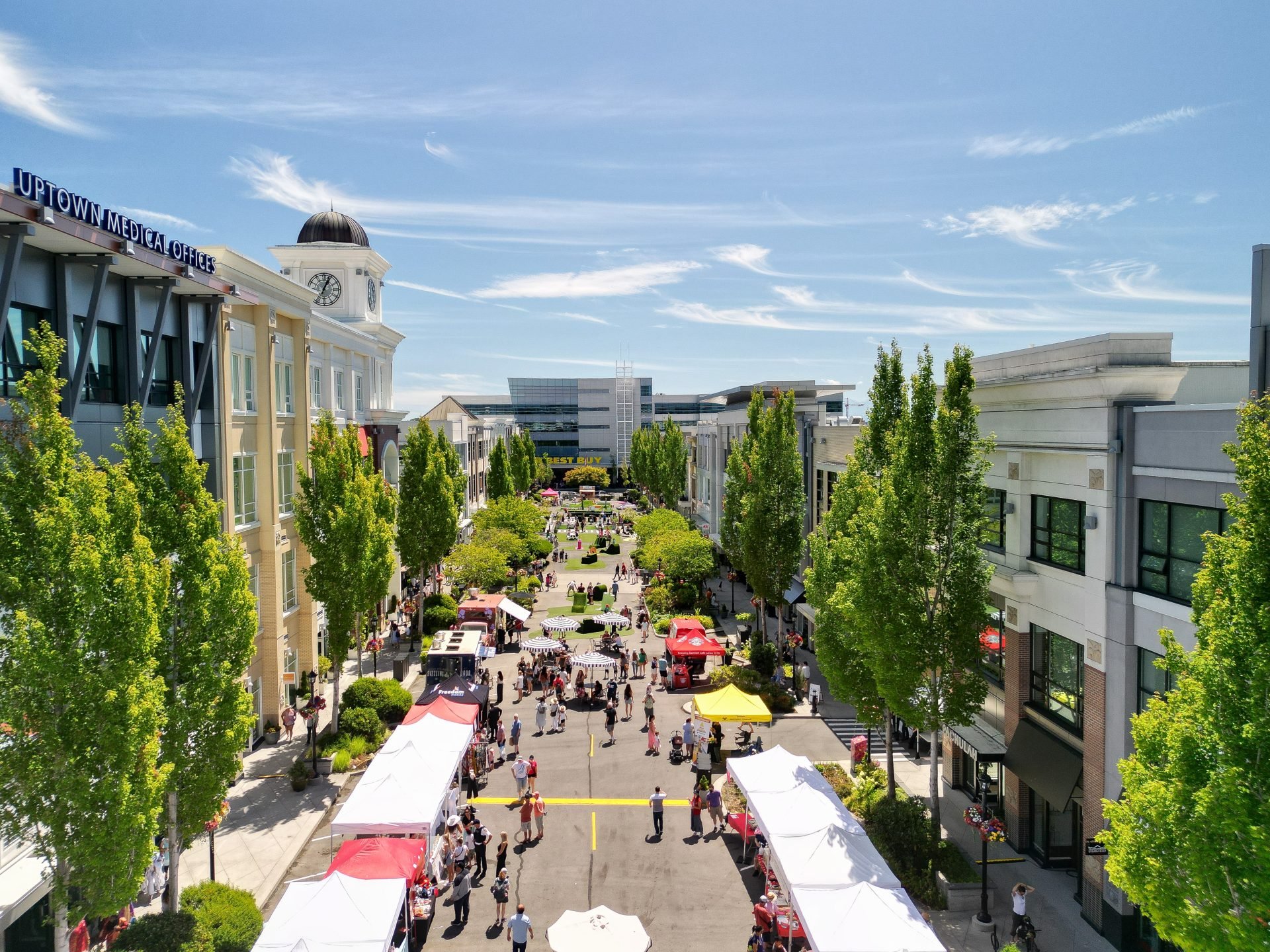 An outdoor shopping and market area with tents and stores, lined with green trees, and a clear blue sky overhead.