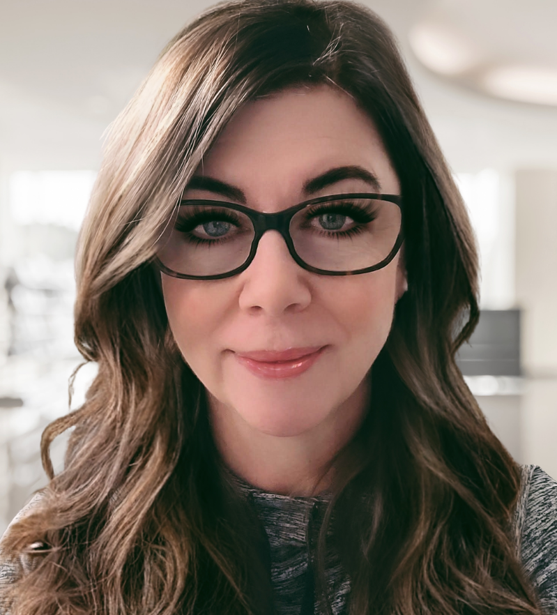A woman with long wavy brown hair wearing black glasses and a gray top, smiling indoors.